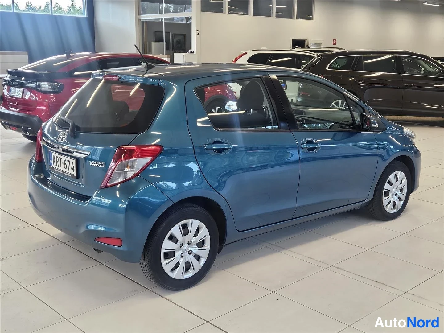 Takaviisto: Sinisenharmaan Toyota Yaris -viistotakakulma autosalissa, rekisterikilpi, takavalot ja vasen kylki näkyvissä.