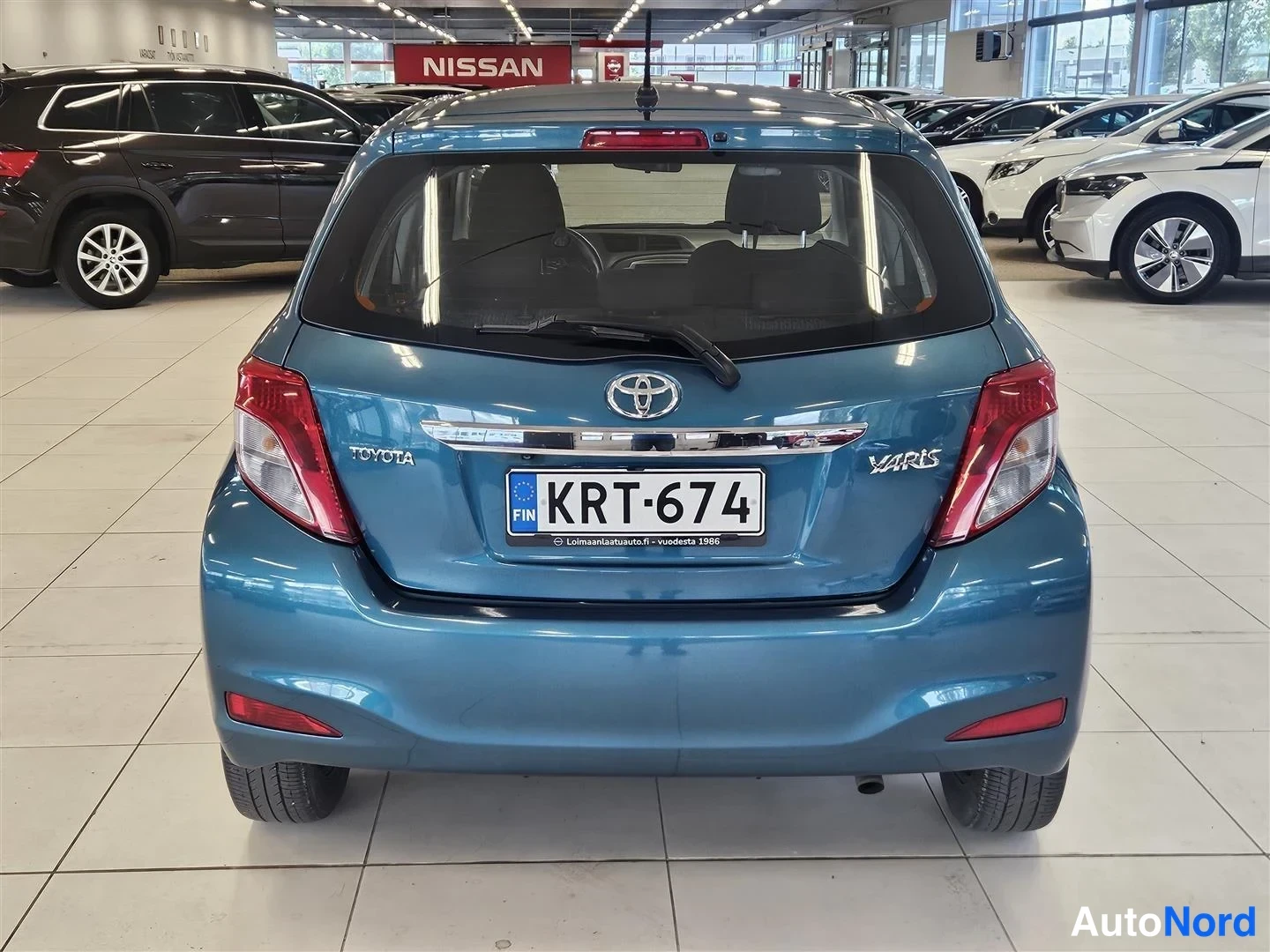 Takaa: Sinisenharmaan Toyota Yarisin takanäkymä autosalissa, rekisterikilpi, TOYOTA- ja YARIS-tekstit takaluukussa.