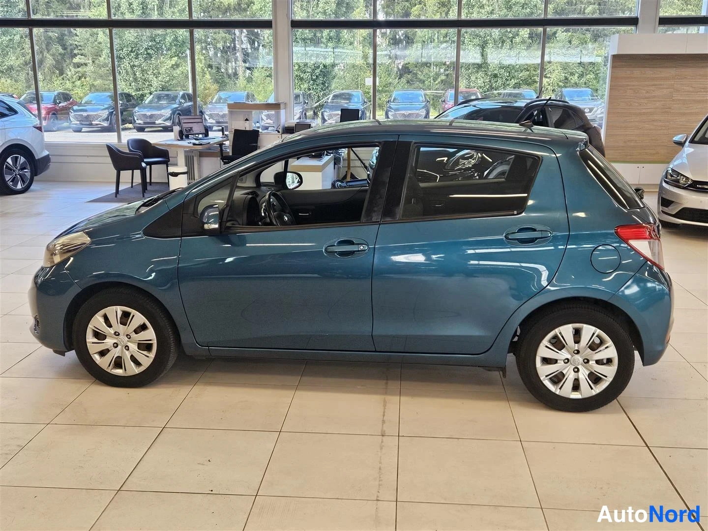 Etuviisto, nokka vasemmalle: Sinisenharmaan Toyota Yarisin sivuseen näkymä autosalissa, vasen kylki ja etuvalo näkyvissä, viisiovinen hatchback.