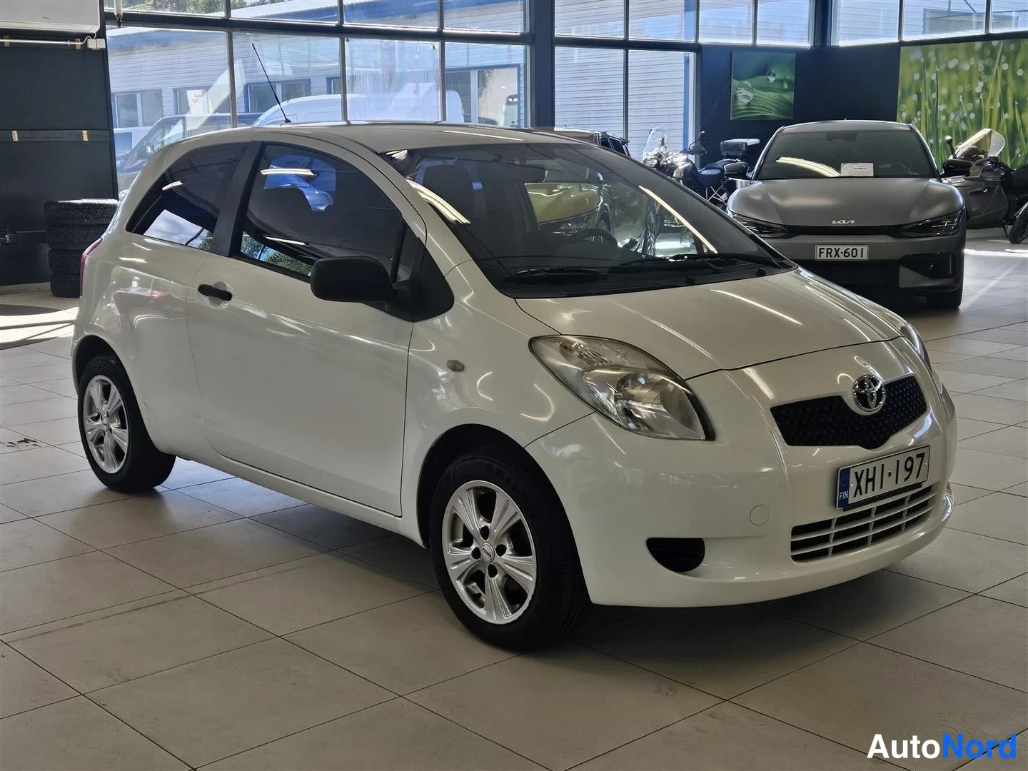 Etuviisto, nokka oikealle: Valkoinen Toyota Yaris -henkilöauto edessä ja sivulta kuvattuna autotilassa, rekisteritunnus.
