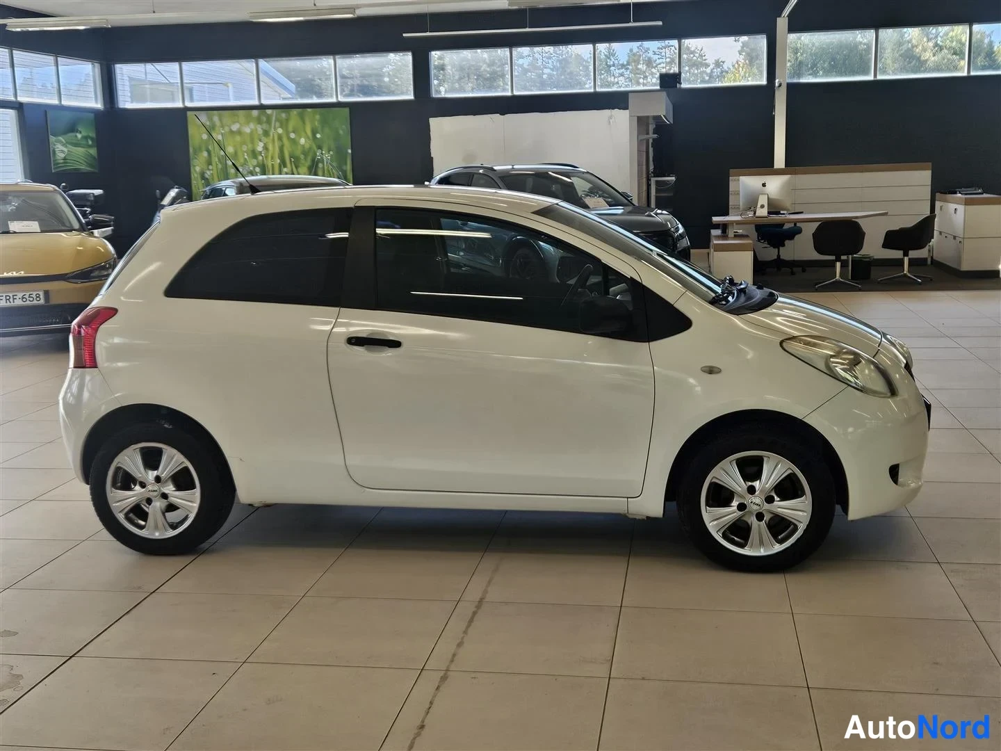 Etuviisto, nokka oikealle: Valkoinen Toyota Yaris -henkilöauto sivulta kuvattuna autotilassa.