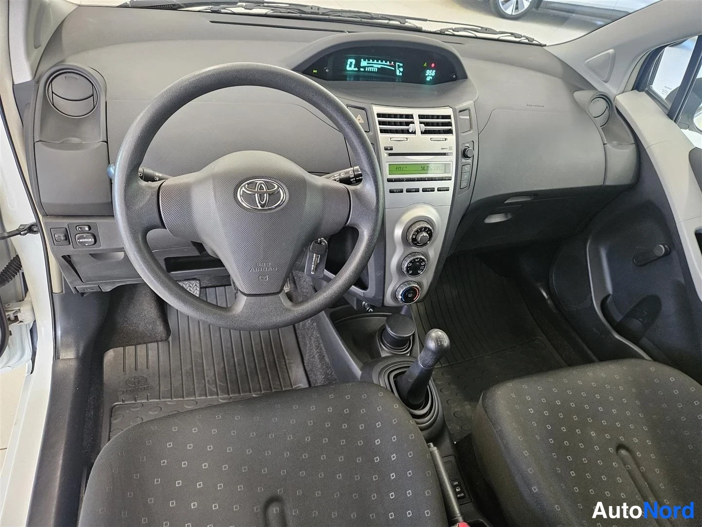 Muu: Toyota Yariksen ohjaamosta etukäsin: Toinen ohjauspyörä Toyota-embleemillä, digitaalinen mittaristo, radio- ja ilmastointilaitteiden säätimet, manuaalivaihteist