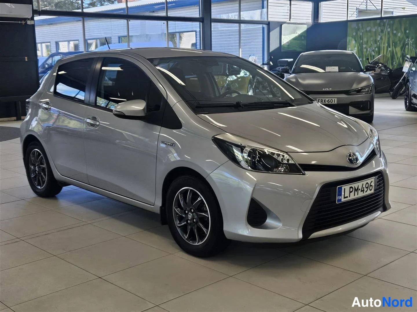 Etuviisto, nokka oikealle: Hopeanharmaa Toyota Yaris viistosti edestä oikealta kuvattuna autosalissa, kyljessä Hybrid-merkintä, rekisteritunnus.
