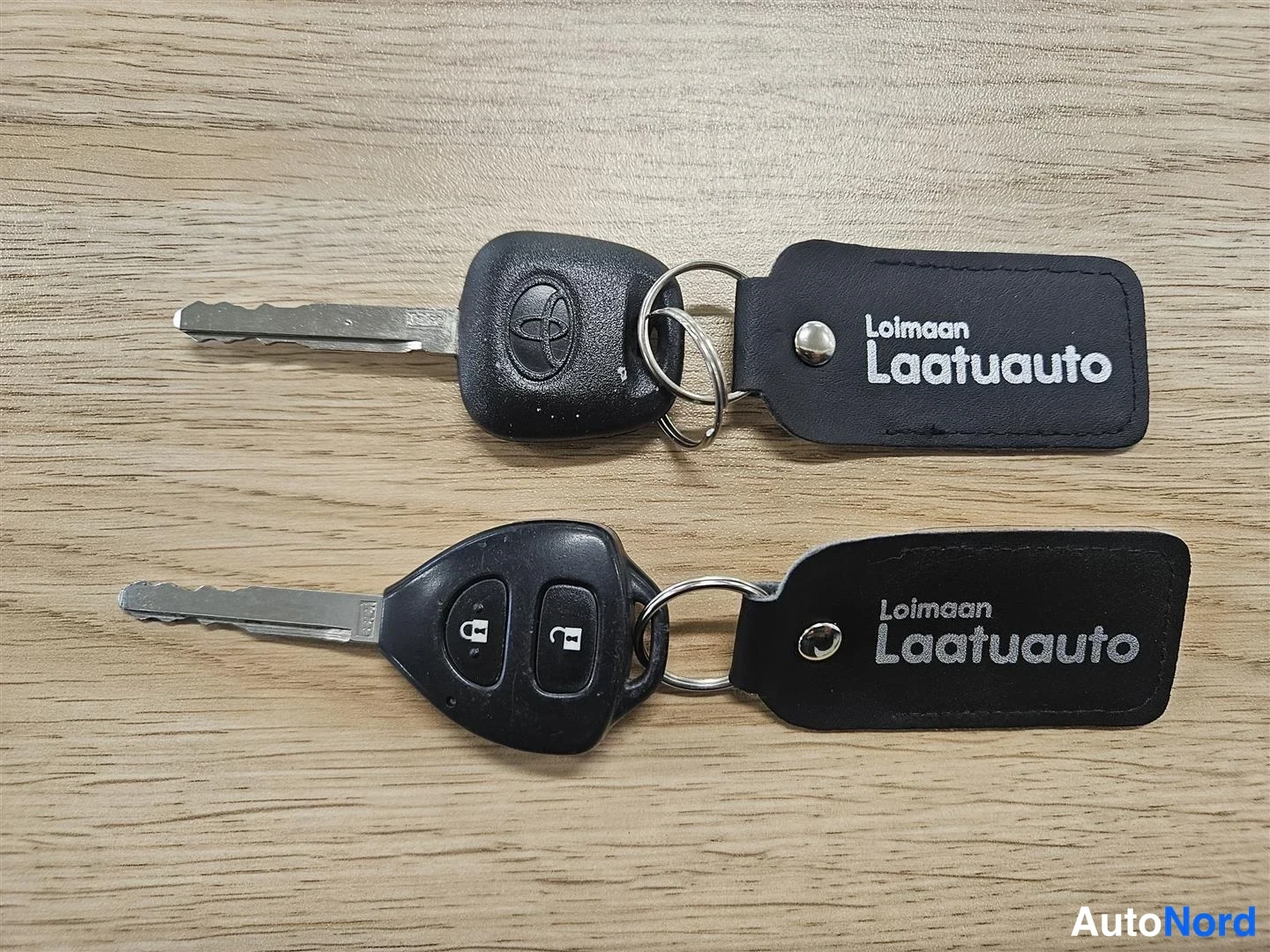 Muu: Kaksi auton avainta mustilla avainnauhoilla, joihin on painettu teksti 'Loimaan Laatuauto', puupohjaisella pinnalla.