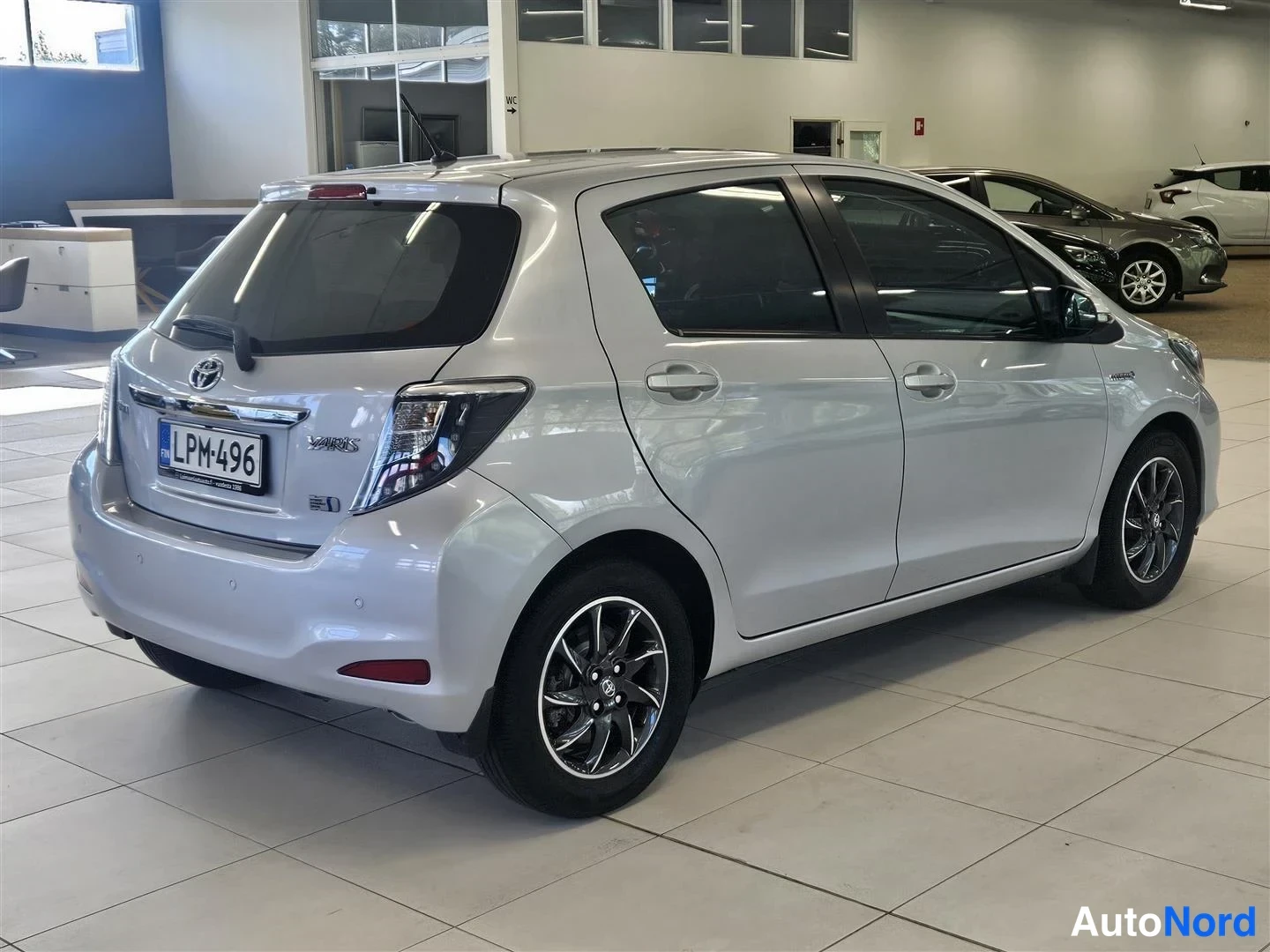 Takaviisto: Hopeanharmaa Toyota Yaris viistosti takaa vasemmalta kuvattuna autosalissa, peräluukussa Yaris-merkintä, rekisteritunnus.