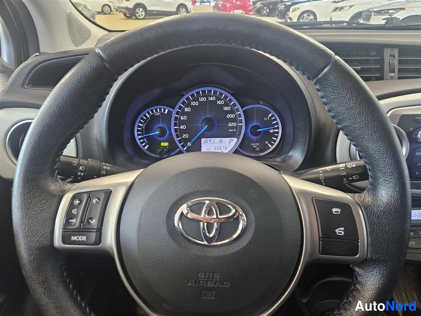 Muu: Ohjaamon näkymä: Toyota-ohjauspyörä ja mittaristo, jossa nopeusmittari, polttoainemittari ja hybridiin viittaava pieni näyttö; matkamittari näyttää lukemaa.
