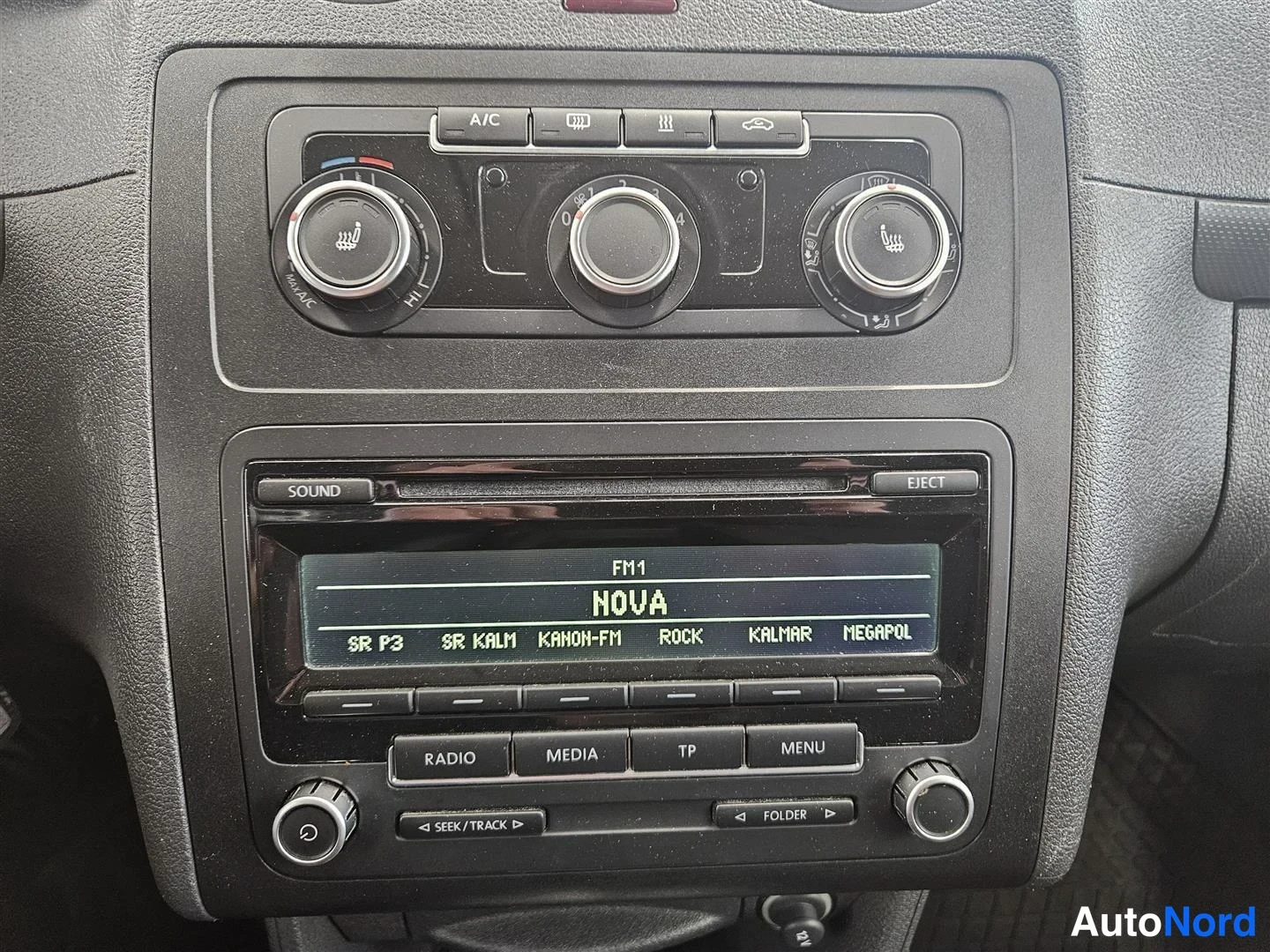 Muu: Kojelaudan keskusta lähikuvassa: ilmastointisäätimet A/C-painikkeella ja radio, jonka näytössä lukee FM1 NOVA sekä kanavalista SR P3, SR KALM, KANON-FM, ROCK, K