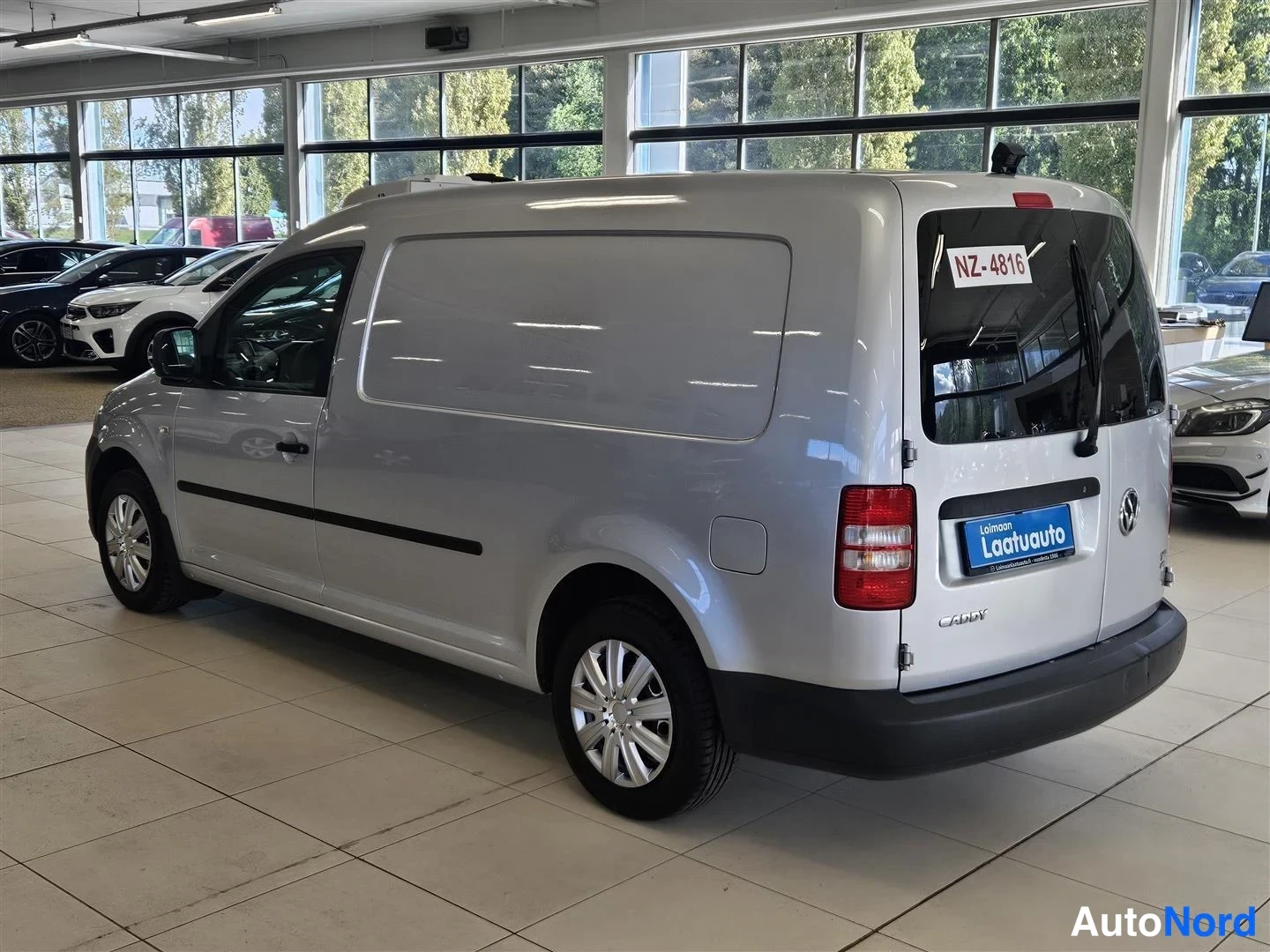 Takaviisto: Pakettiauton takakulma oikealta: hopeinen kylki ja takaosa, NZ-4816 -tarra takalasissa, musta alakorkki, autosalin ikkunaseinä taustalla.