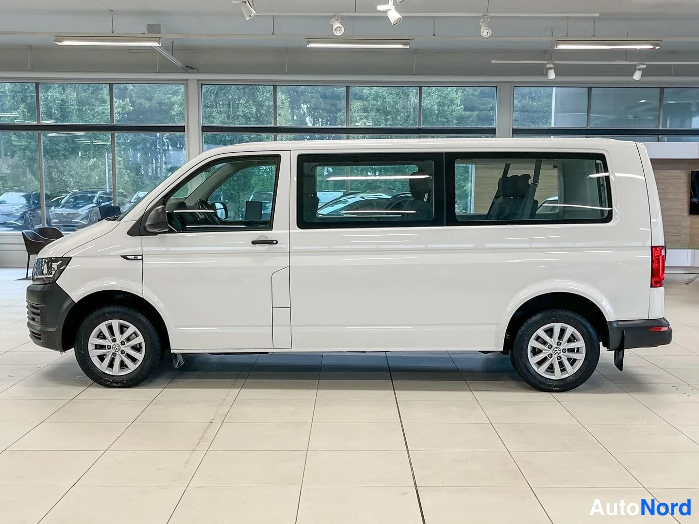 Takaviisto: Sivukulmasta kuvattu valkoinen Volkswagen Transporter -pakettiauto näyttelyn lattialla; pitkä sivulinja, perävalot näkyvät oikealla reunalla.
