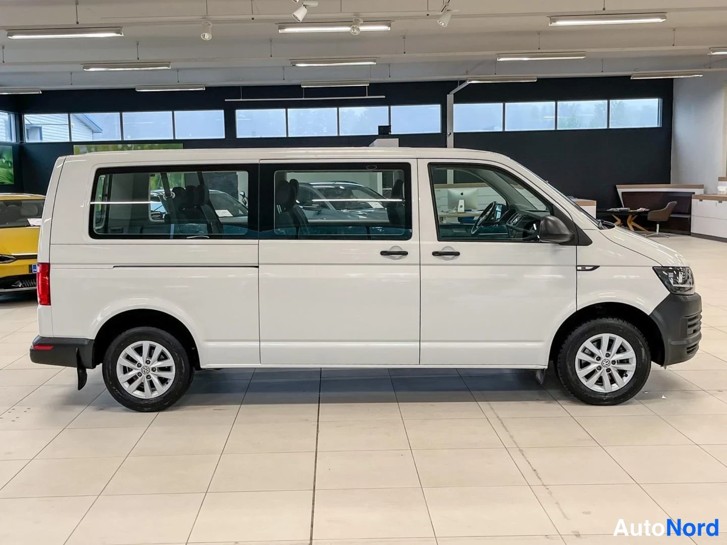 Etuviisto, nokka oikealle: Valkoinen Volkswagen Transporter -pakettiauto sivulta vasemmasta nokasta kuvattuna; etuvalo ja tumma etupuskuri näkyvät kuvan oikeassa reunassa.