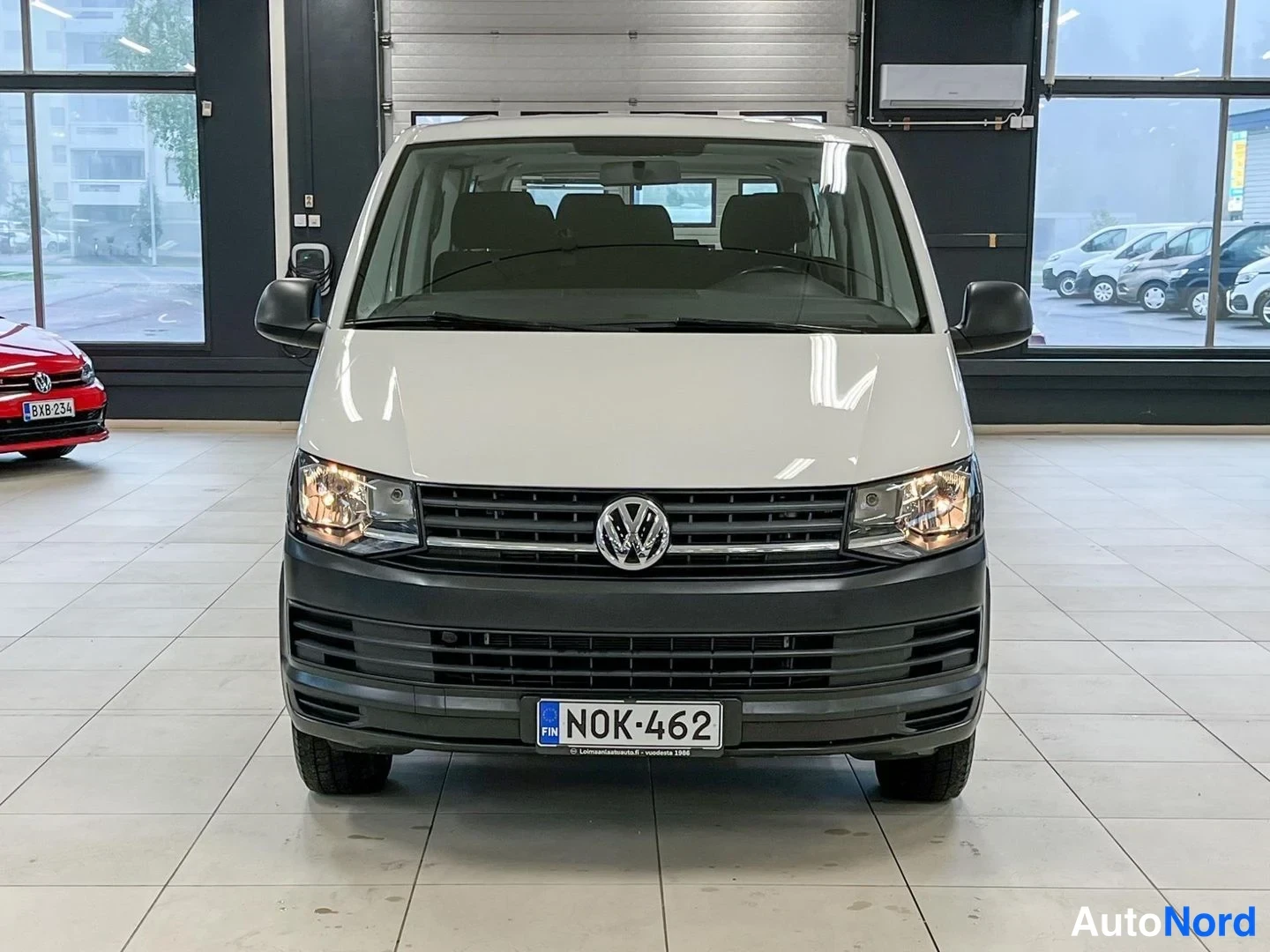 Edestä: Valkoinen Volkswagen Transporter -pakettiauto suoraan edestäpäin; ajovalot päällä, rekisterikilpi, tumma etupuskuri ja Volkswagen-tunnus konepellissä.