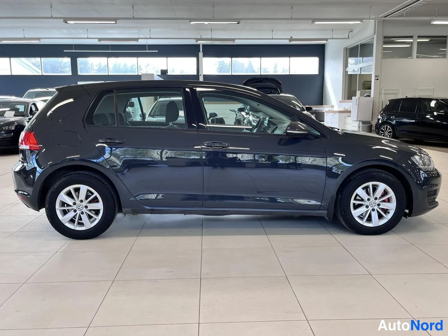 Sivulta: Tummanharmaan viisiovisen hatchbackin sivuprofiili showroomissa, hopeavanteet molemmissa pyörissä.