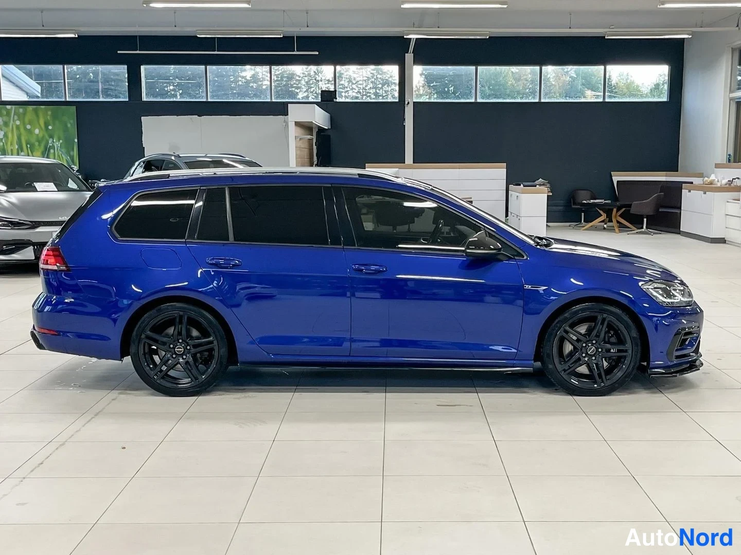 Sivulta: Sininen farmariauto sivusta oikealta kuvattuna, mustat pyörät ja tummat ikkunat näkyvät, autosalin sisätiloissa.