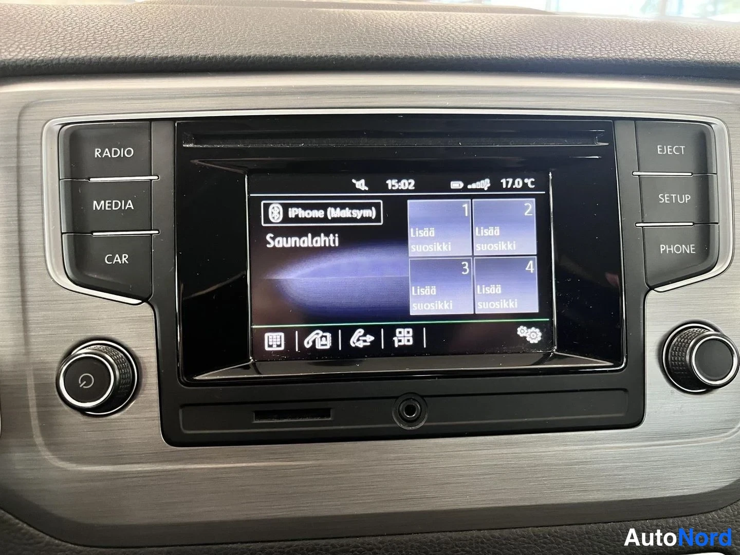 Sivulta: Autojen radiopaneeli lähikuvassa: näytöllä iPhone (Maksym), Saunalahti ja lisää suosikki -painikkeet, 15:02 ja 17,0 °C ylhäällä, RADIO/MEDIA/CAR vasemmalla ja E