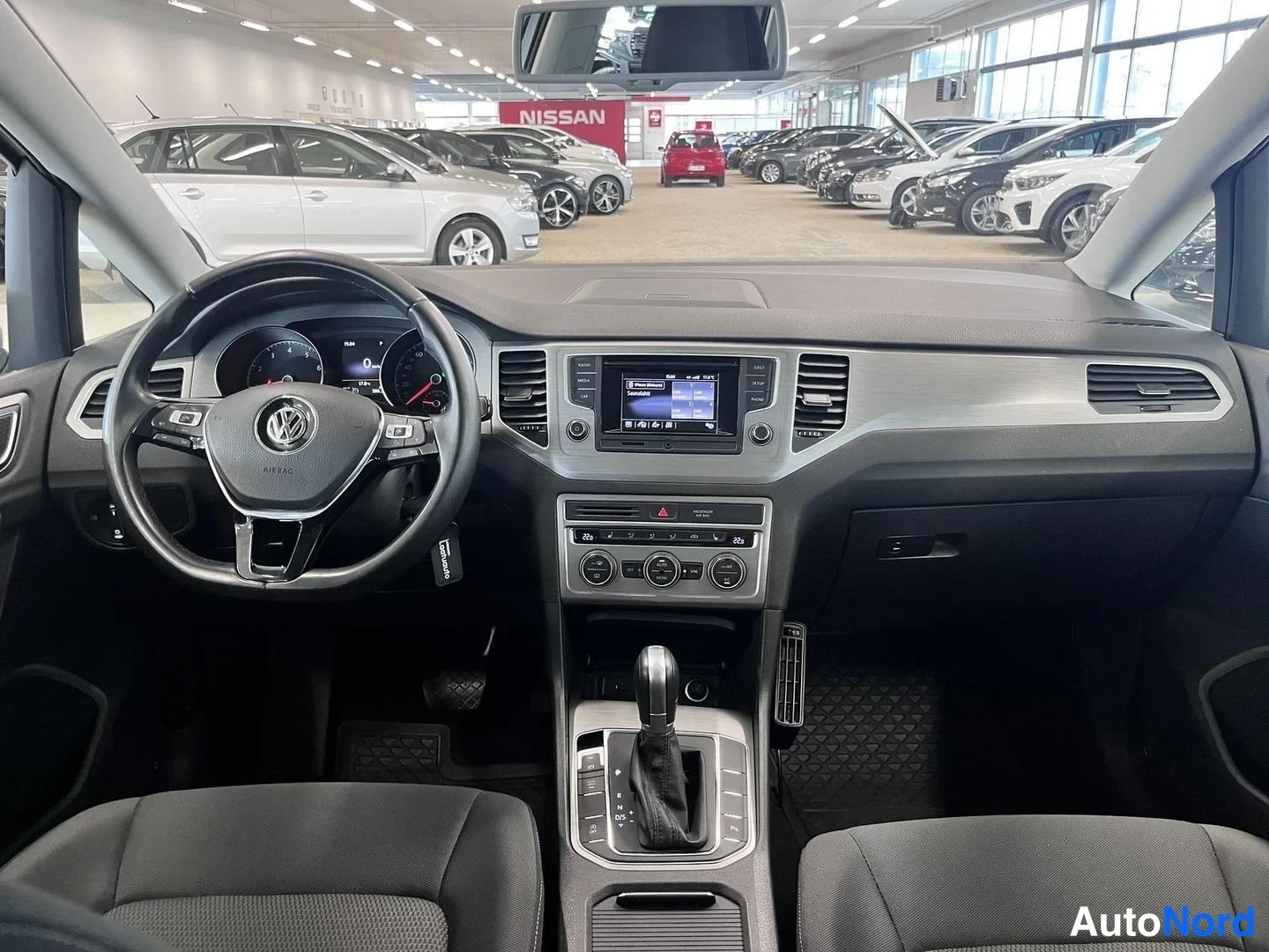 Muu: Auton ohjaamo autonmyyntihallissa: ohjauspyörä VW-merkillä, kojelauta, radio/kosketusnäyttö, ilmastoinnin säätimet ja automaattivaihteen valitsin keskikonsoliss