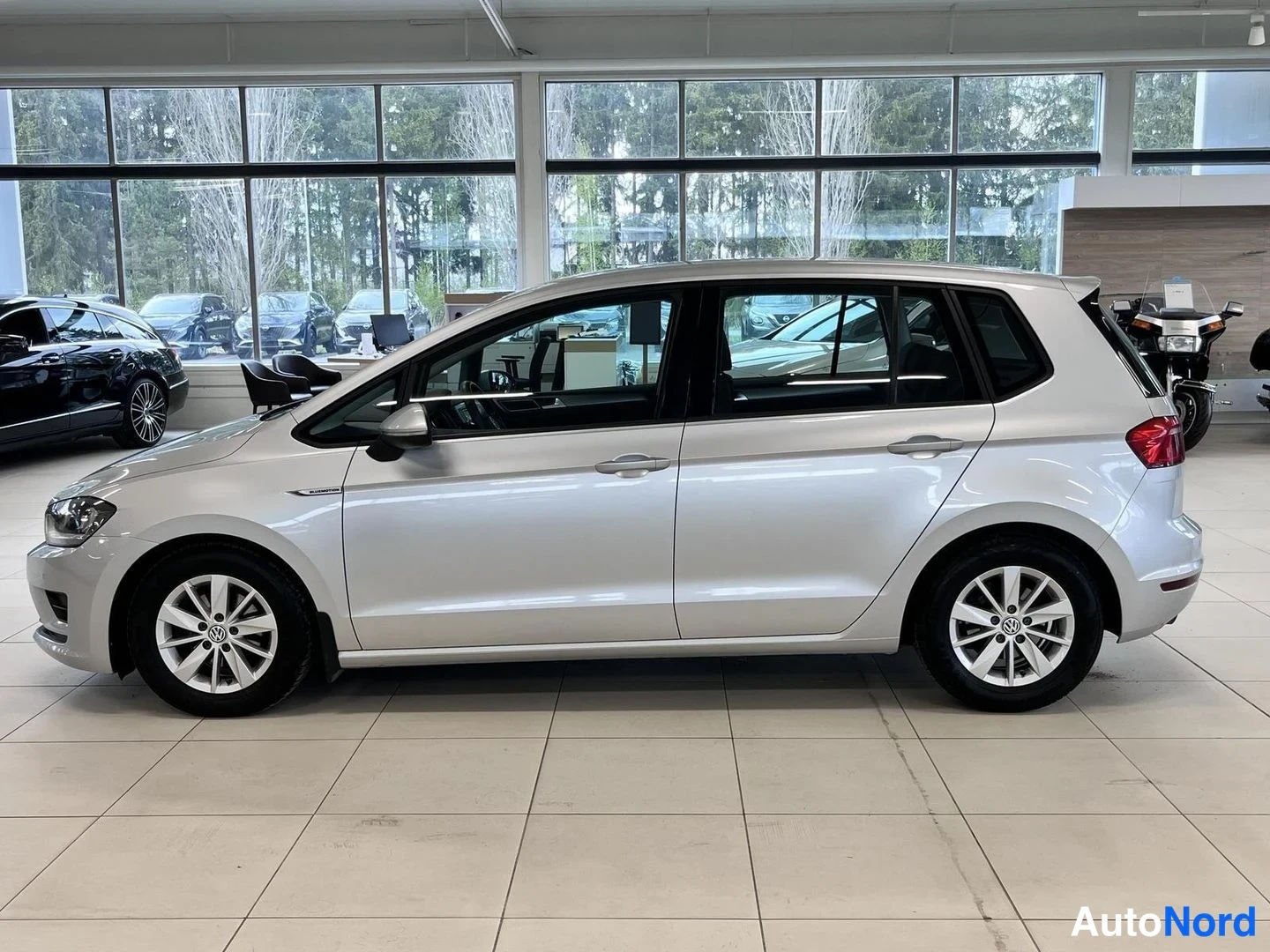 Etuviisto, nokka vasemmalle: Hopeanvärinen Volkswagen Golf Sportsvan vasemmasta sivusta etuviistoon kuvattuna autosalissa.