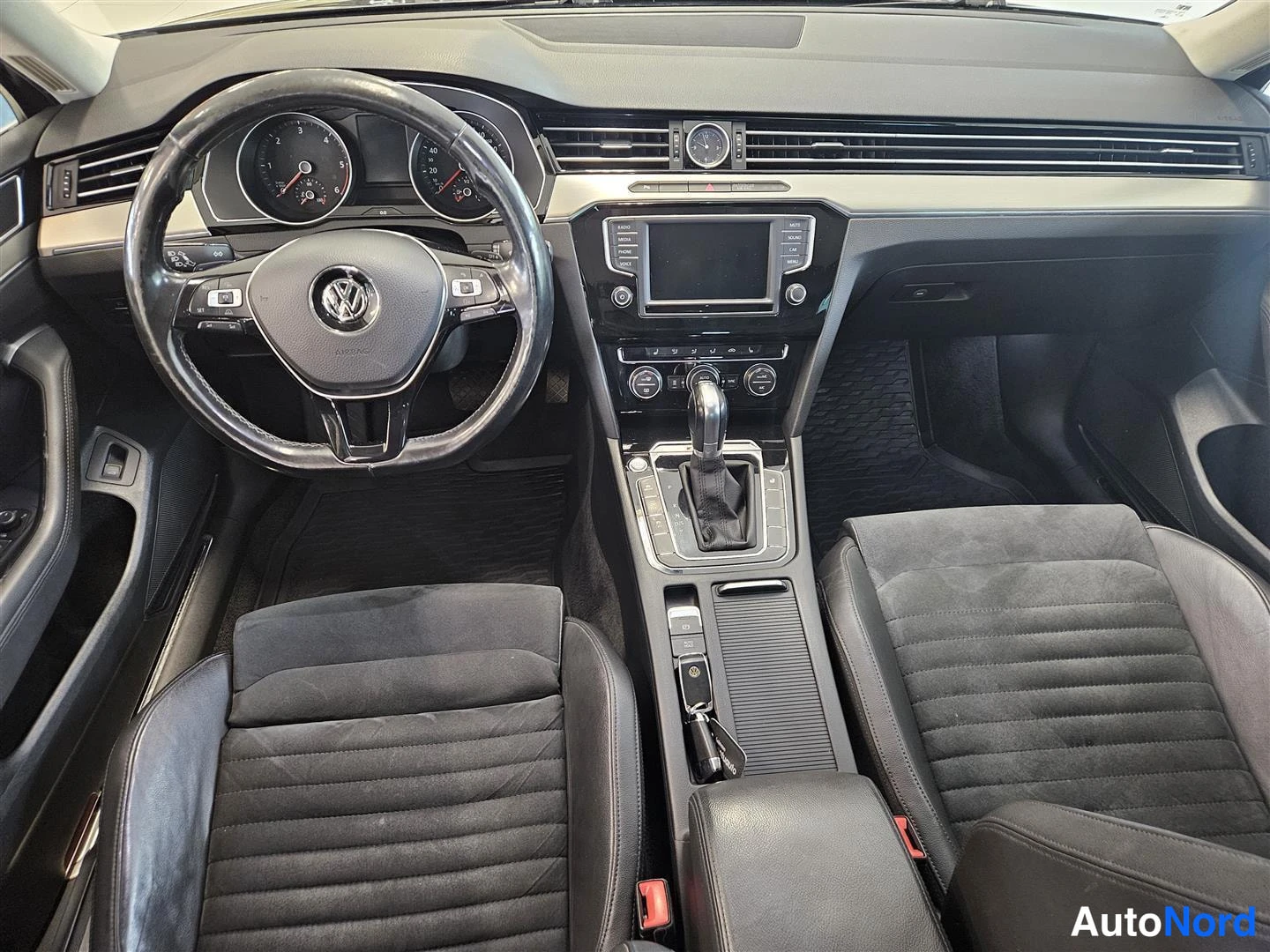Muu: Kuljettajan näkökulma kojelautaan: VW-ohjauspyörä, mittaristo, kosketusnäyttö ja automaattivaihteiston valitsin, tummat etuistuimet.
