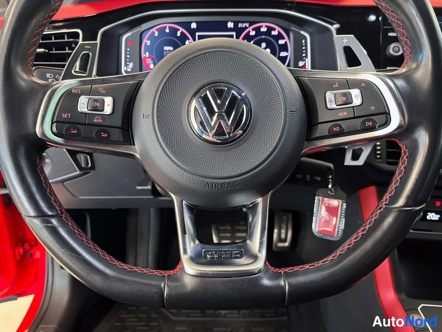 Muu: Lähikuva VW-ohjauspyörästä punaisilla ompeleilla ja AIRBAG-merkinnällä, taustalla digitaalinen mittaristo.