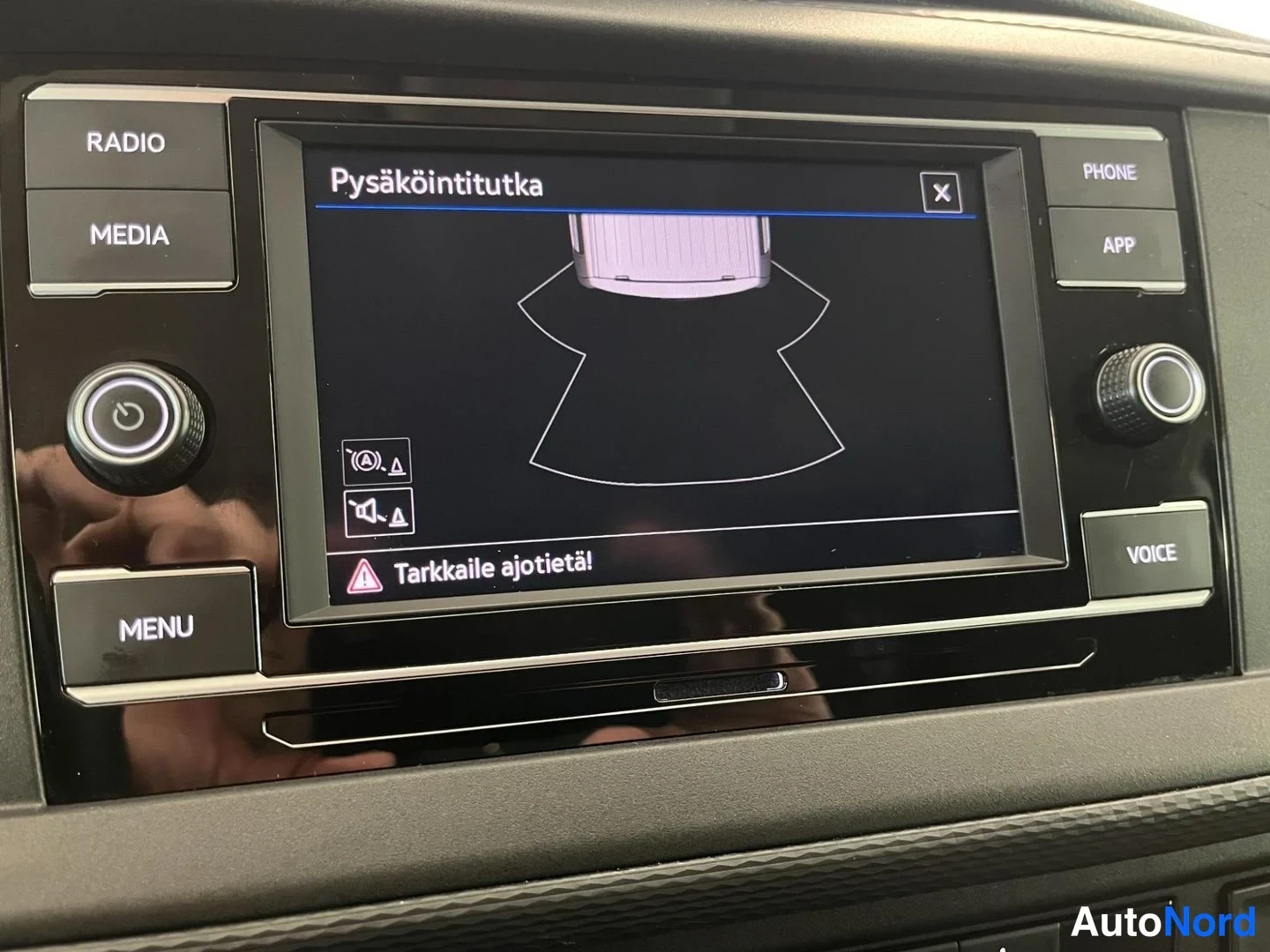 Yksityiskohta: Radioyksikön näyttö näyttää Pysäköintitutka-näkymän auton apunäytöllä, alareunassa varoitus 'Tarkkaile ajotietä!'.
