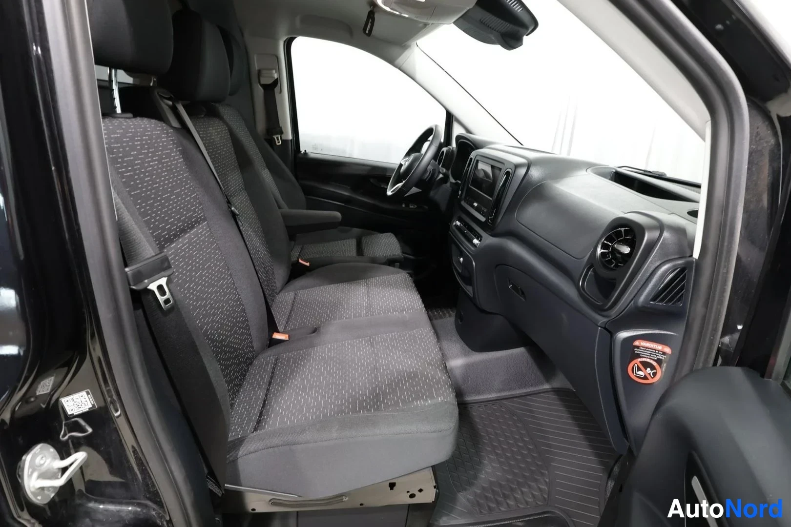 Muu: Sisätilakuva Mercedes-Benz Vito -auton matkustamosta takaapäin: tummat kankaisistuimet kaksi riviä ja etuistuimet avonaisesta liukuovesta kuvattuna.