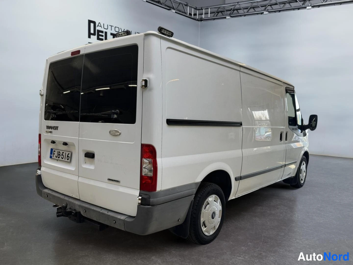 Takaviisto: Valkoisen Ford Transit -pakettiauton takaosa ja oikea kylki kolmannesviistossa, takarenkaiden kotelot ja perävaunukytkin näkyvät.