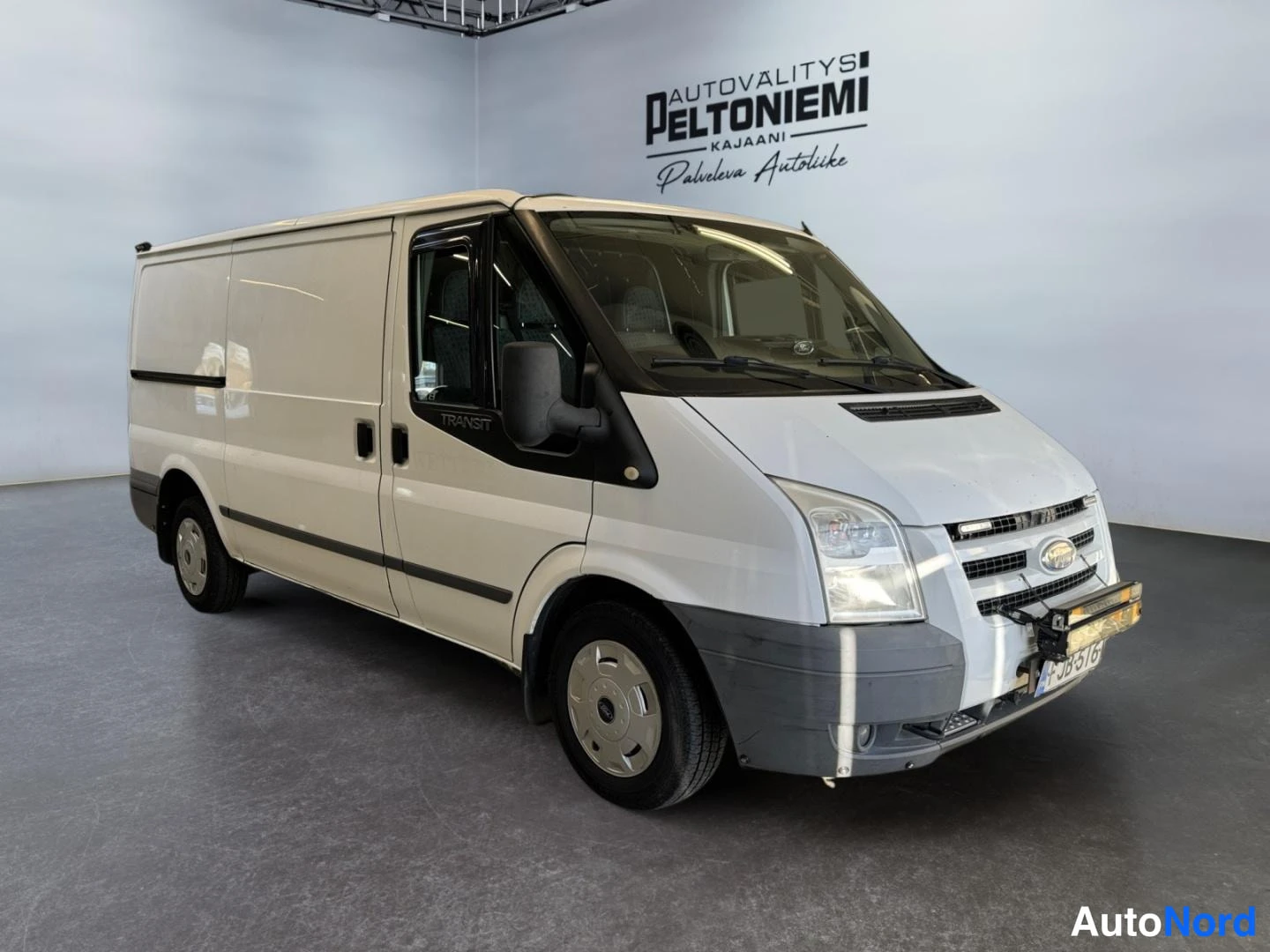 Etuviisto, nokka oikealle: Ford Transit -pakettiauto etuviistosta oikealta kuvattuna autoliikkeen tiloissa, vaaleanvalkoinen koripanelit, tummat ikkunakehykset, rekisterikilpi näky