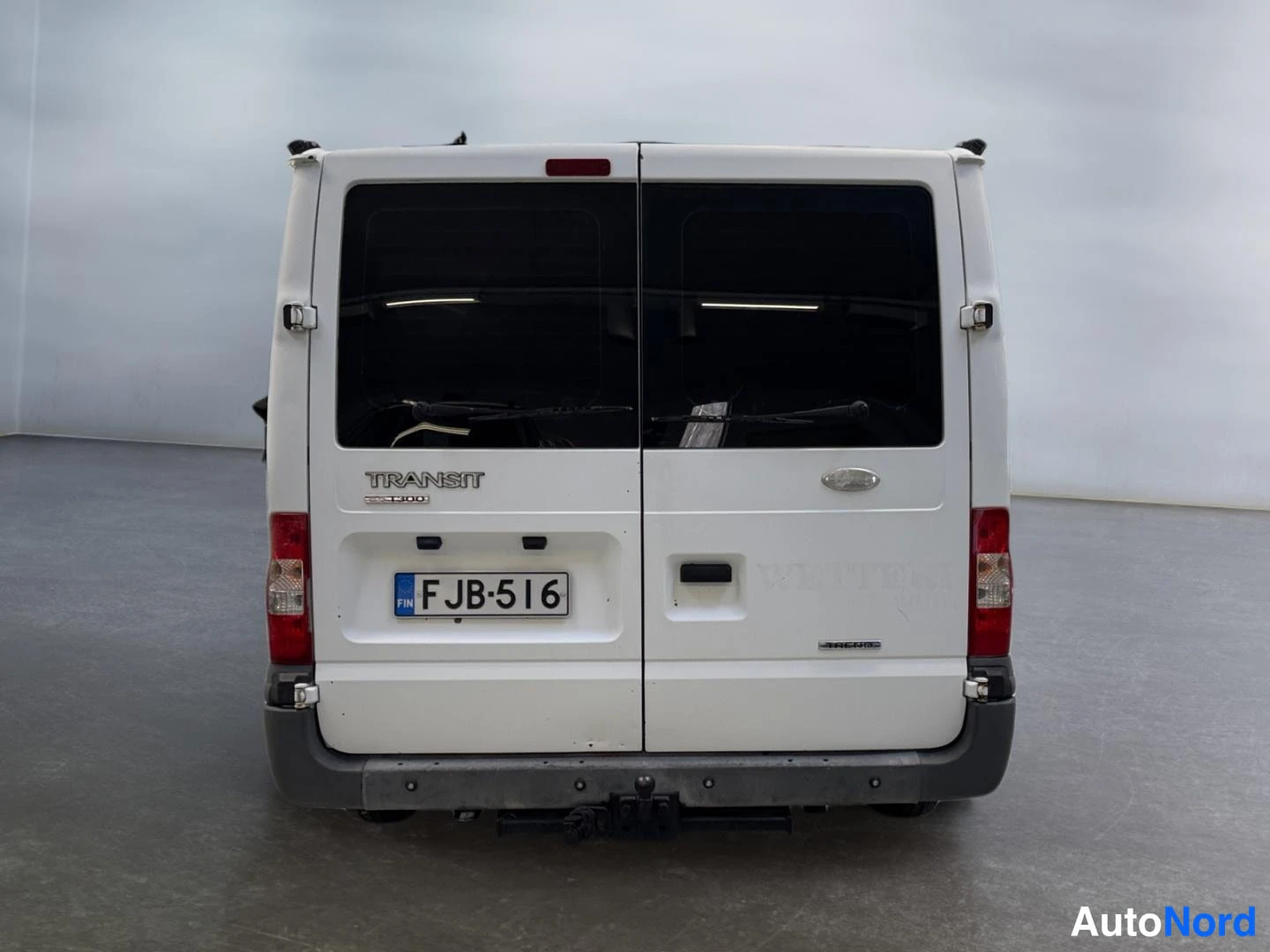Takaa: Valkoisen Ford Transit -pakettiauton takaosa suoraan takaa, rekisterikilpi ja TRANSIT-teksti takaluukussa.