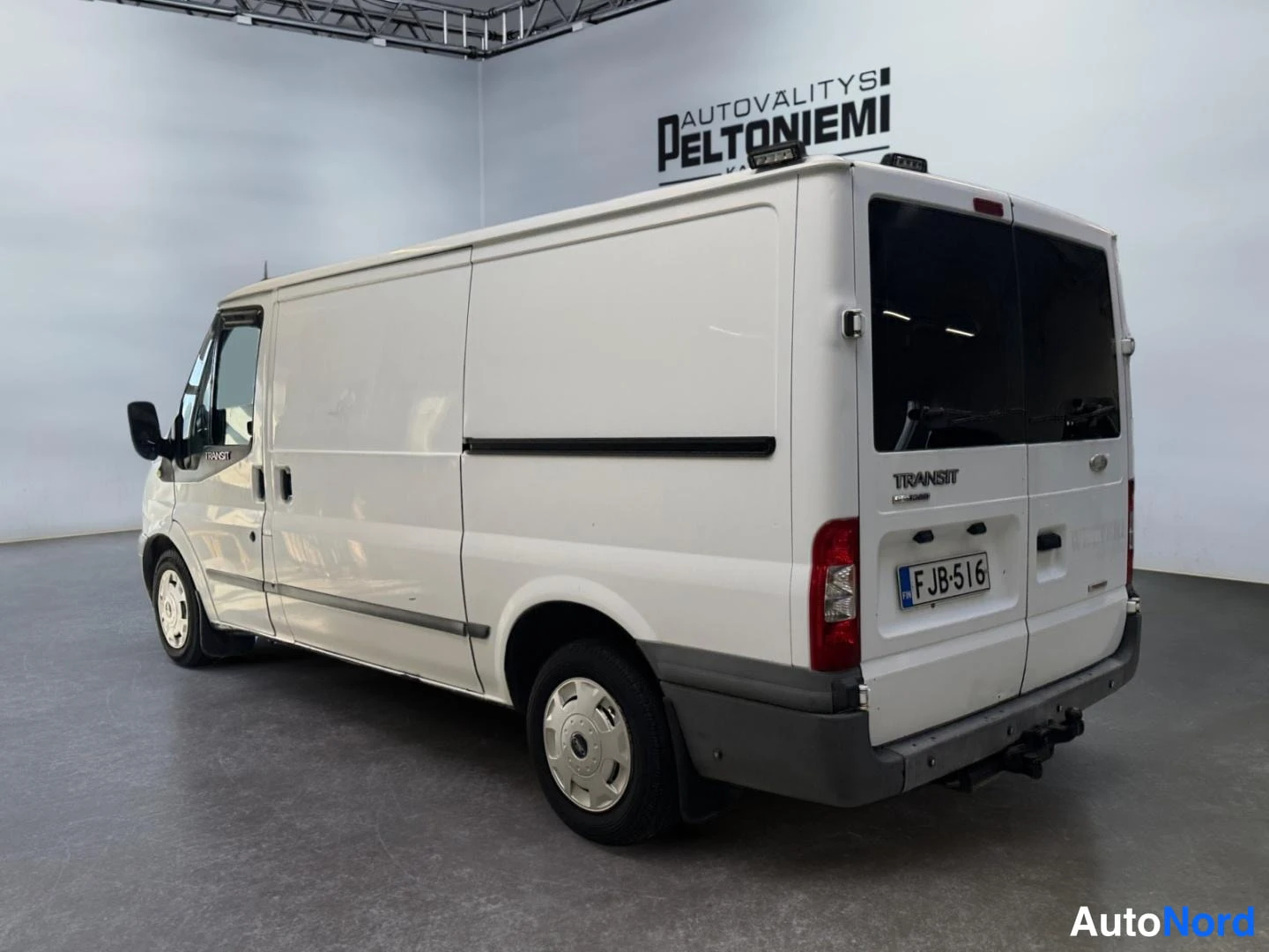 Takaviisto: Valkoinen Ford Transit -pakettiauto takaosa ja vasen kylki kolmannesviistossa sisätiloissa, rekisterikilpi ja TRANSIT-teksti takaluukussa.