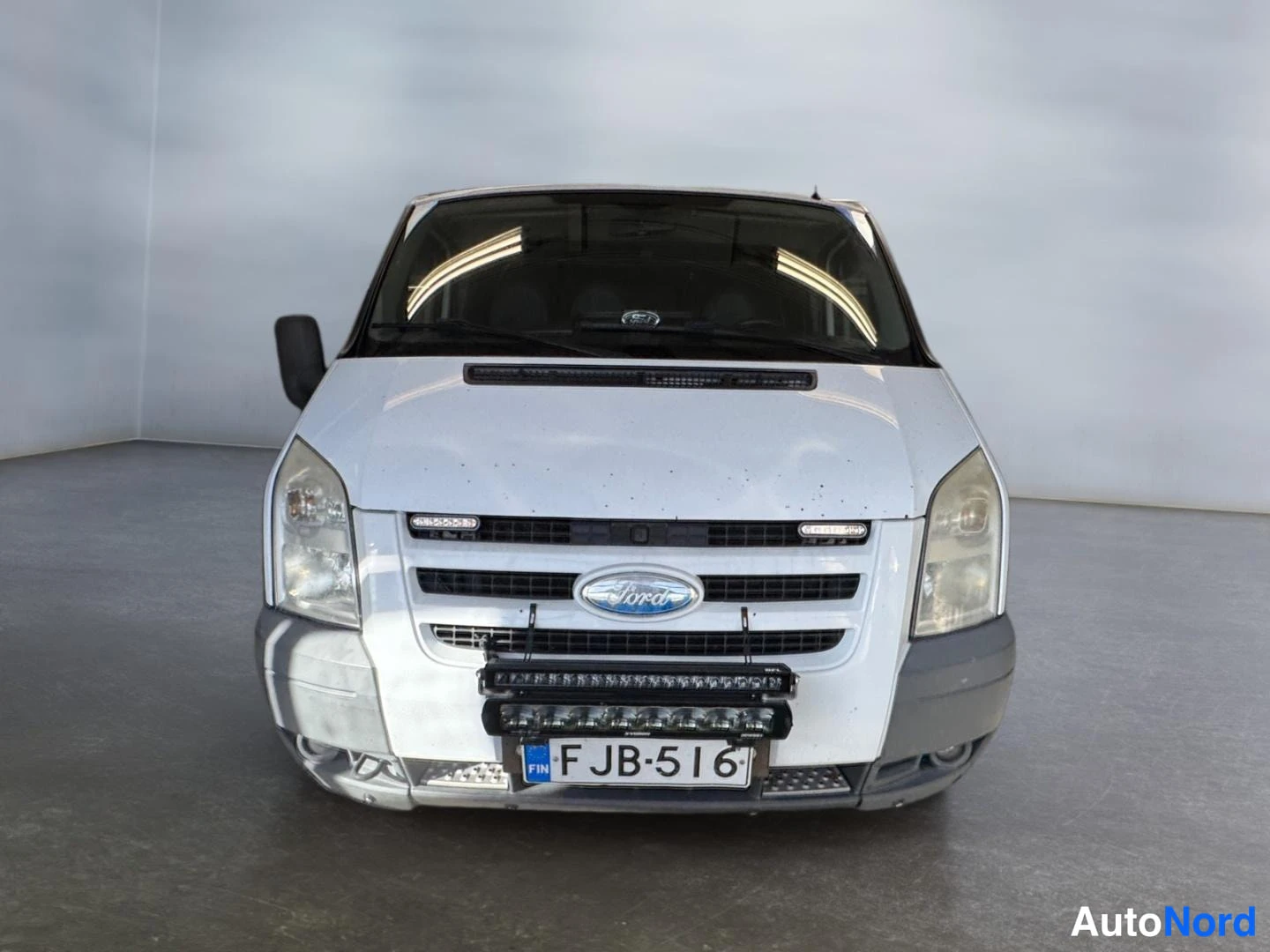 Edestä: Suora etänäkymä valkoisen Ford Transitin keulasta; jäähdyttimen säleikössä Ford-merkki, puskurissa lisävalokisko ja rekisterikilpi.