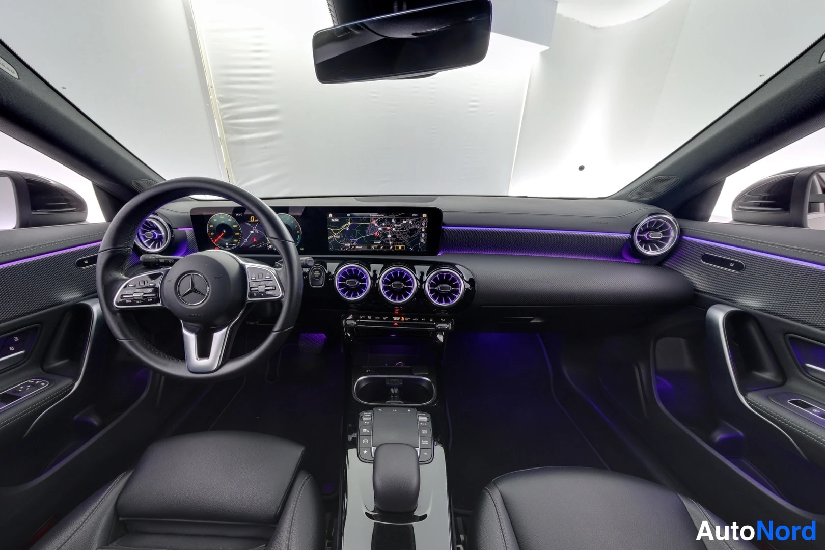 Edestä: Mercedes-Benz CLA:n ohjaamo edestä kuvattuna: ohjauspyörä, digitaalinen mittaristo, keskikonsolin näyttö navigointikartalla, violetit korostusvalot ja mustat is