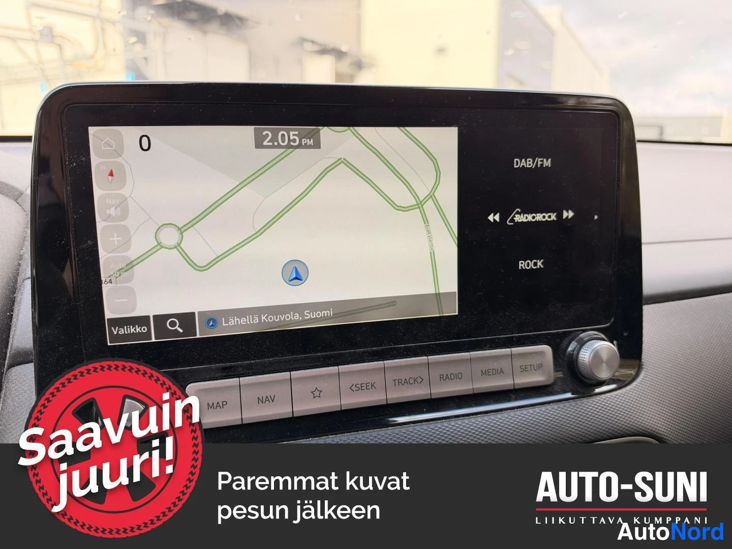 Yksityiskohta: Auton kojelautaan integroitu infotainment-näyttö, jolla näkyy navigointikartta tekstillä 'Lähellä Kouvola, Suomi' sekä DAB/FM-radioasetus Radio Rock. Näytön all