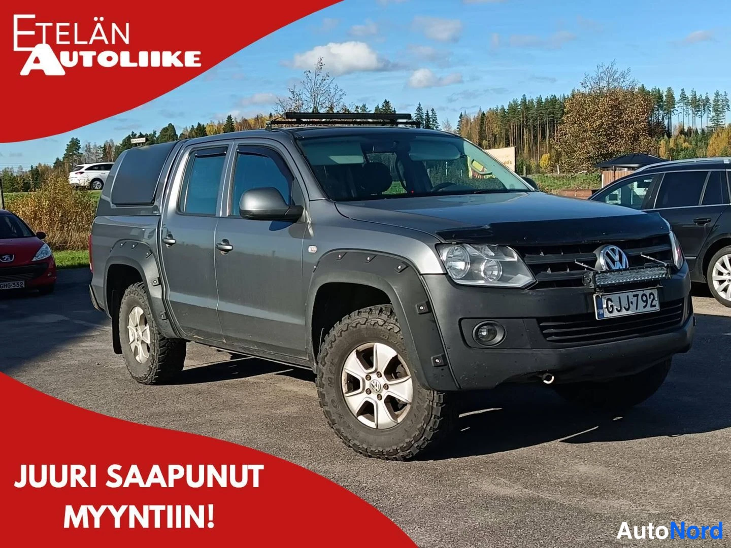 Etuviisto, nokka oikealle: Harmaa Volkswagen Amarok -nappakuormaaja vinosti edestä vasemmalta kuvattuna, tummat levylevikkeet, LED-kevytpalkki konepellin etureunassa ja kattoteline