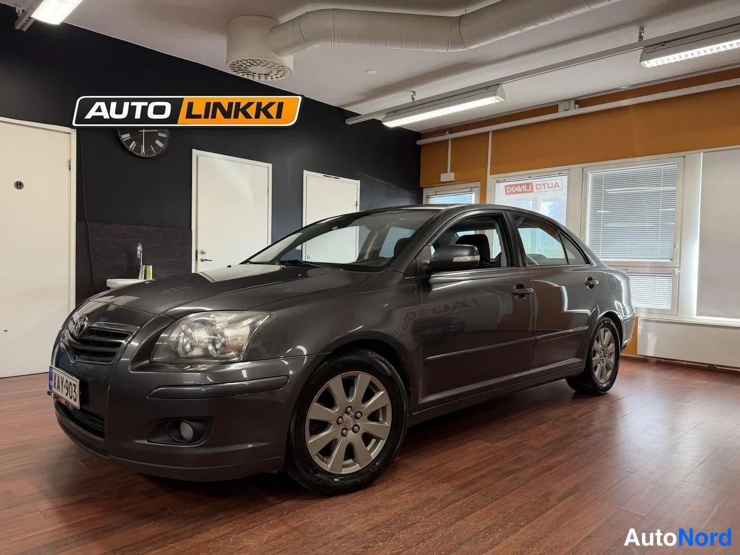Etuviisto, nokka vasemmalle: Tummanharmaa Toyota Avensis -sedan esillä sisätilassa, etukulmasta oikealta; etugrilli, ajovalot ja oikea sivu näkyvät.