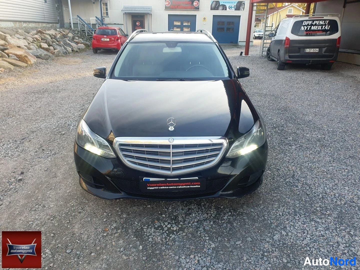 Edestä: Mustan farmariautin keula suoraan edestä päin; etusäleikkö, Mercedes-tähti ja etuvalot näkyvät, etuvalot palavat.