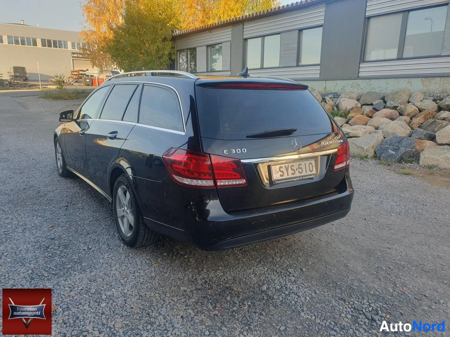 Takaviisto: Musta Mercedes-Benz E 300 BlueTec Hybrid -farmari taka-kulmanurkasta, takavalo ja benz-tähti konepellin keskellä, rekisterikilpi ''.