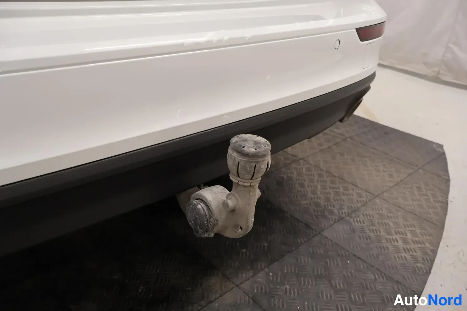 Takaa: Auton takaosassa kiinnitetty irrotettava vetokoukku, jossa pyöreä sähköliitin. Auto on valkoinen ja seisoo mustalla kenkälevykodeilla päällystetyllä alustalla.