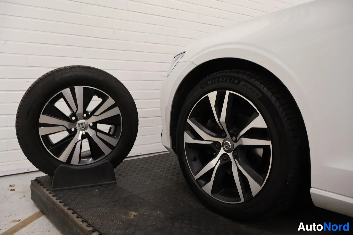 Edestä: Valkoisen auton etupyörä ja alumiinivanne lähikuvassa, vieressä varapyörä telineessä Michelin-renkaalla.