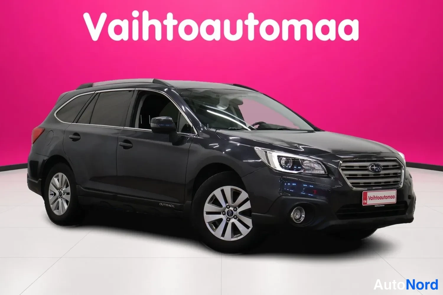 Etuviisto, nokka oikealle: Tummanharmaan farmariauto Subarun etunurkka kuvattuna oikealta, valkopohjaiset vanteet ja kattokaaret näkyvät, tausta vaaleanpunainen.