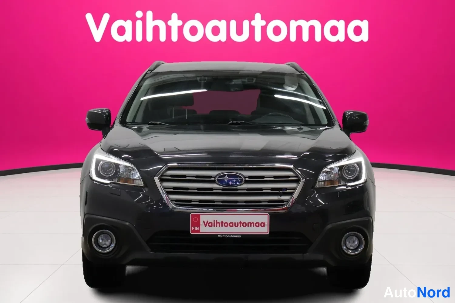 Edestä: Tummanharmaan farmariauton Suubrun keula suoraan edestä, maski ja Subaru-merkki keskilta, sumuvalot pohjassa, tausta vaaleanpunainen.