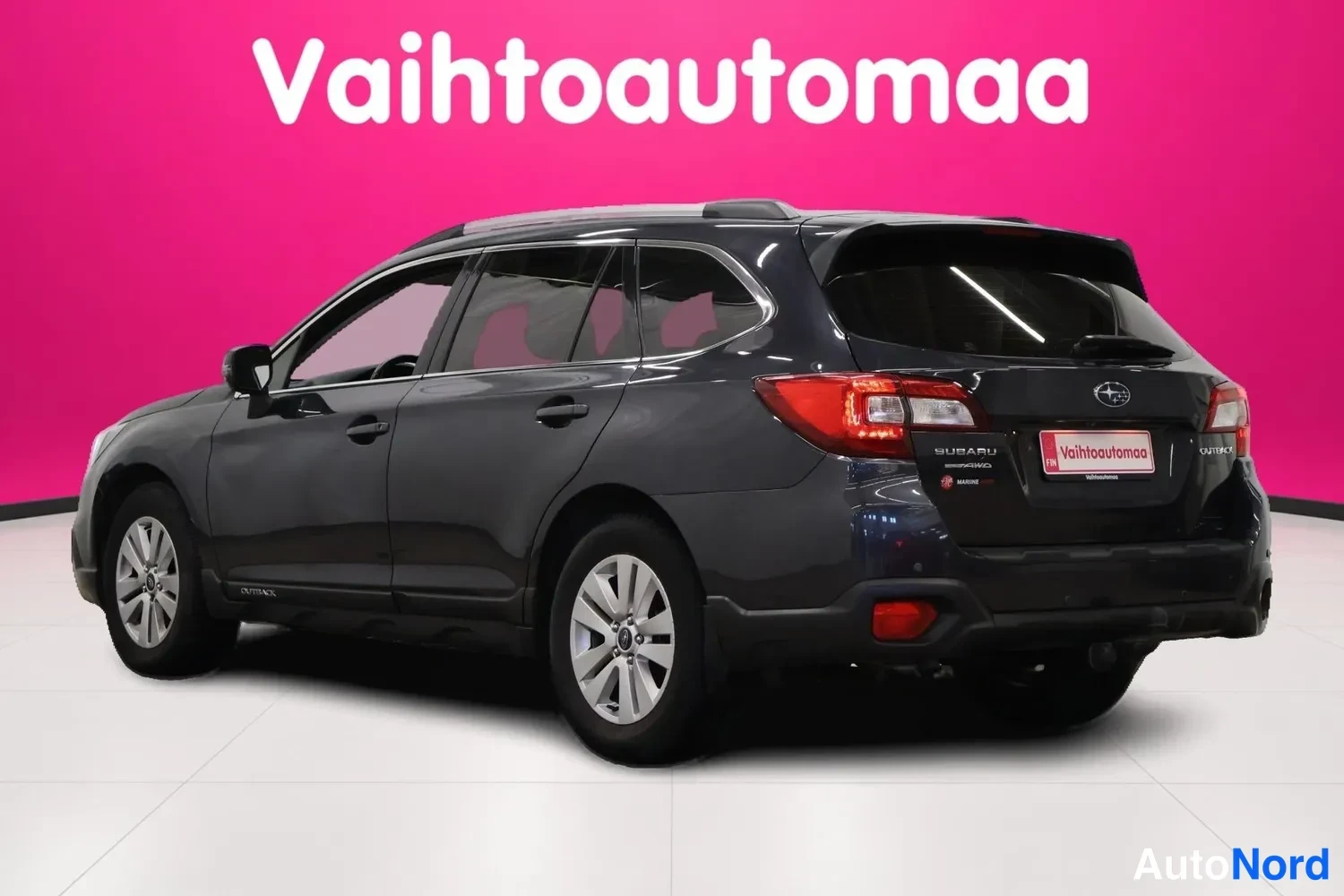 Takaviisto: Tummanharmaan farmariauton Subarun takakulma kuvattuna vasemmalta, takavalot ja Subaru-merkki rekisterikilven yllä, tausta vaaleanpunainen.