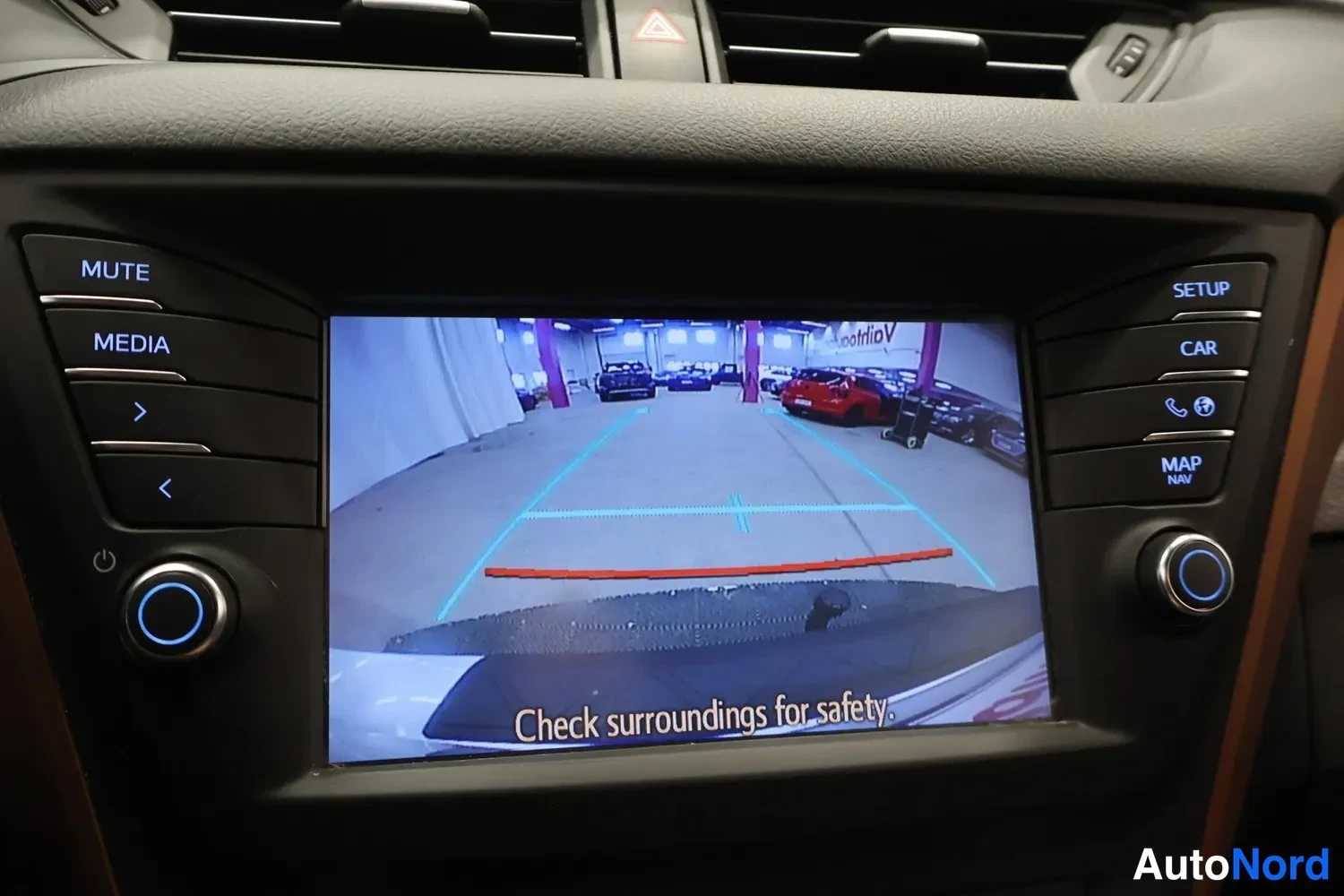 Muu: Kojelaudan infotainment-näyttö näyttää peruutuskameran kuvan ohjausviivoineen ja tekstillä 'Check surroundings for safety'.