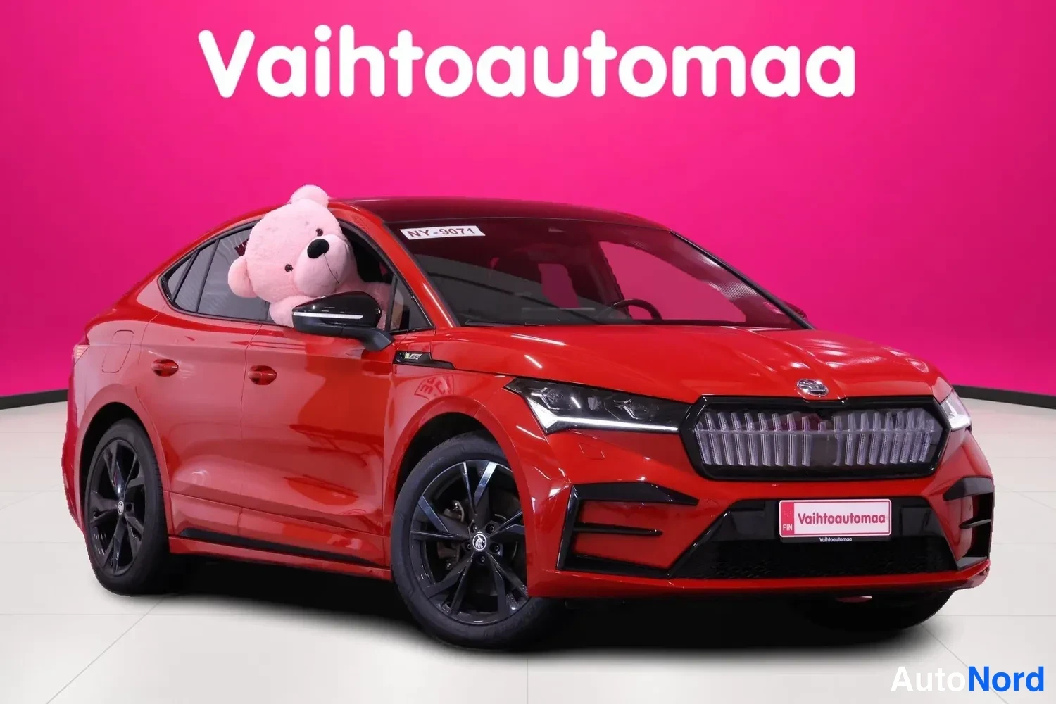 Etuviisto, nokka oikealle: Punainen henkilöauto etuviistosta, etugrin ja pyörät näkyvät; pehmolelu takana.