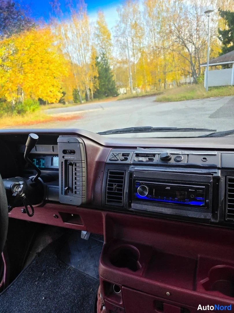 Muu: Kojelaudan lähikuva: JVC-radio sinisellä valaistuksella, ilmastointisuuttimet, kupitelineet ja tummin nahka korostettuna.