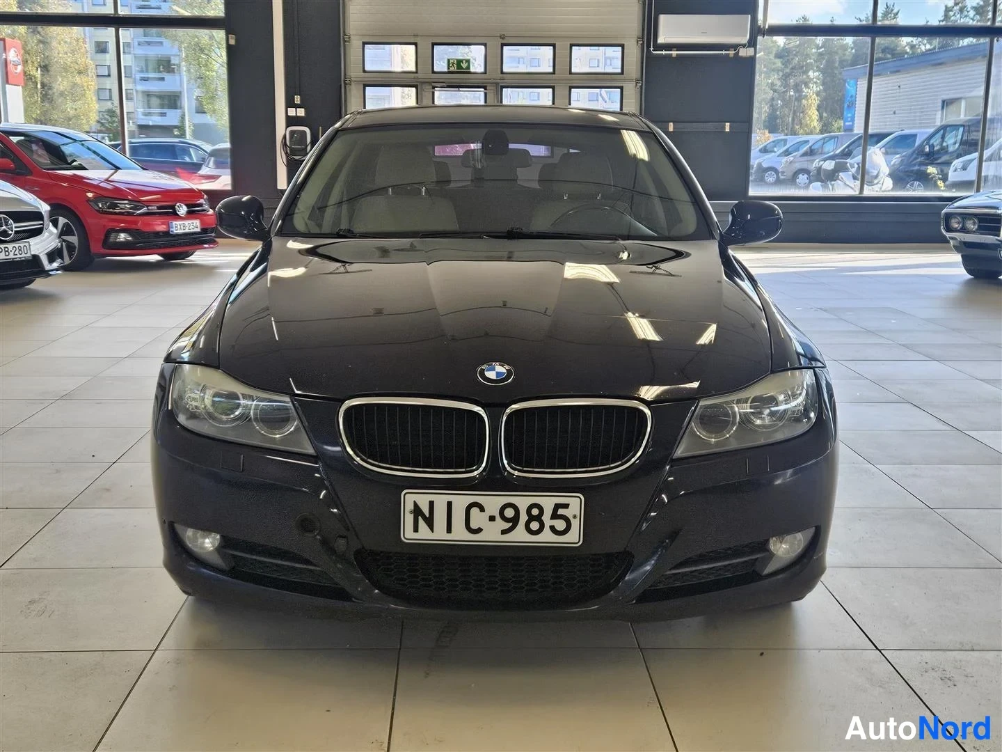 Edestä: Mustan BMW 318:n suora etunäkymä, konepellin keskusosa kiiltää showroomin valaistuksessa, rekisterikilpi.