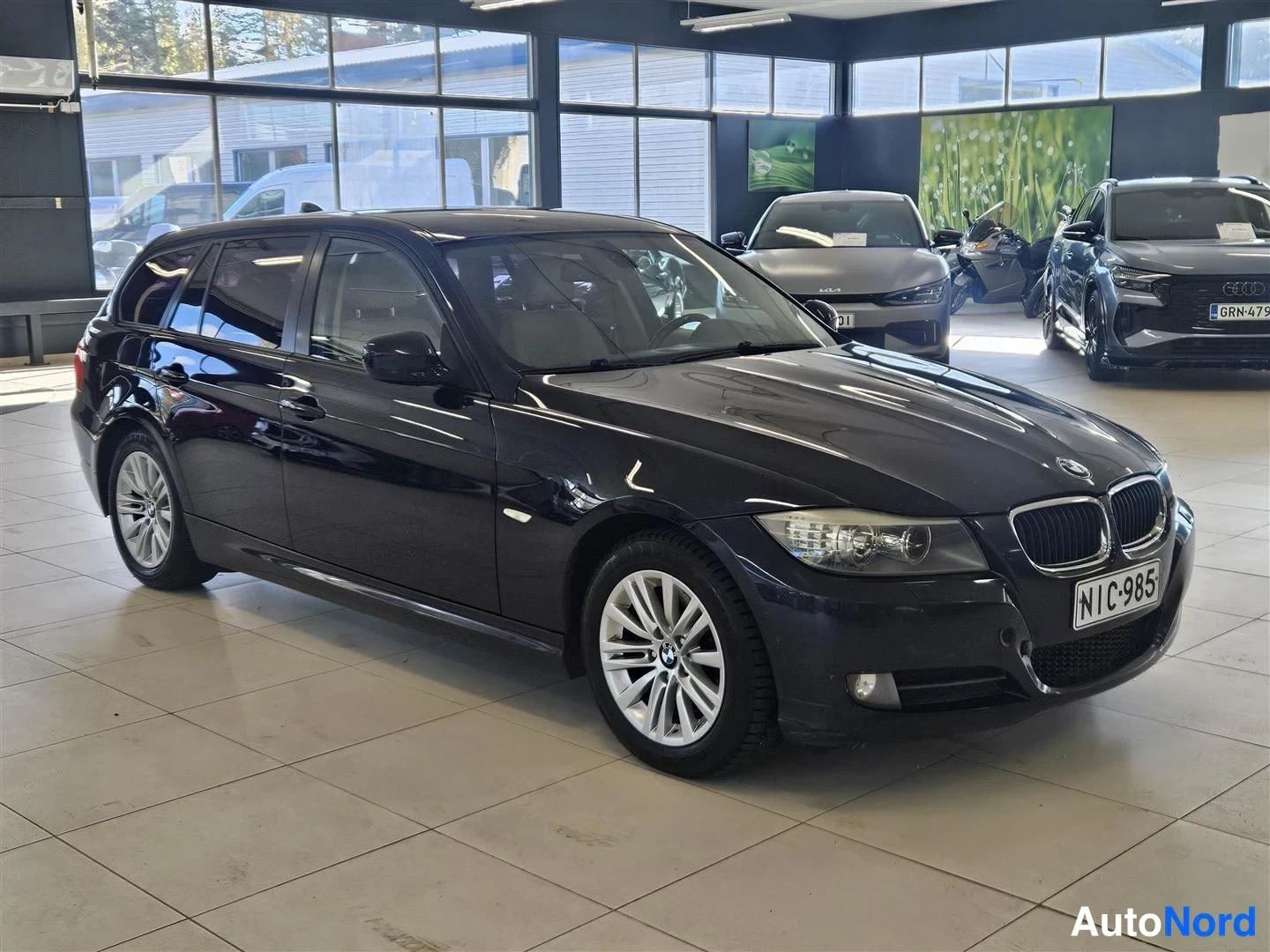 Etuviisto, nokka oikealle: Musta BMW 318 farmari etukulmasta oikealta, rekisterikilpi, taustalla muita ajoneuvoja esityshallissa.