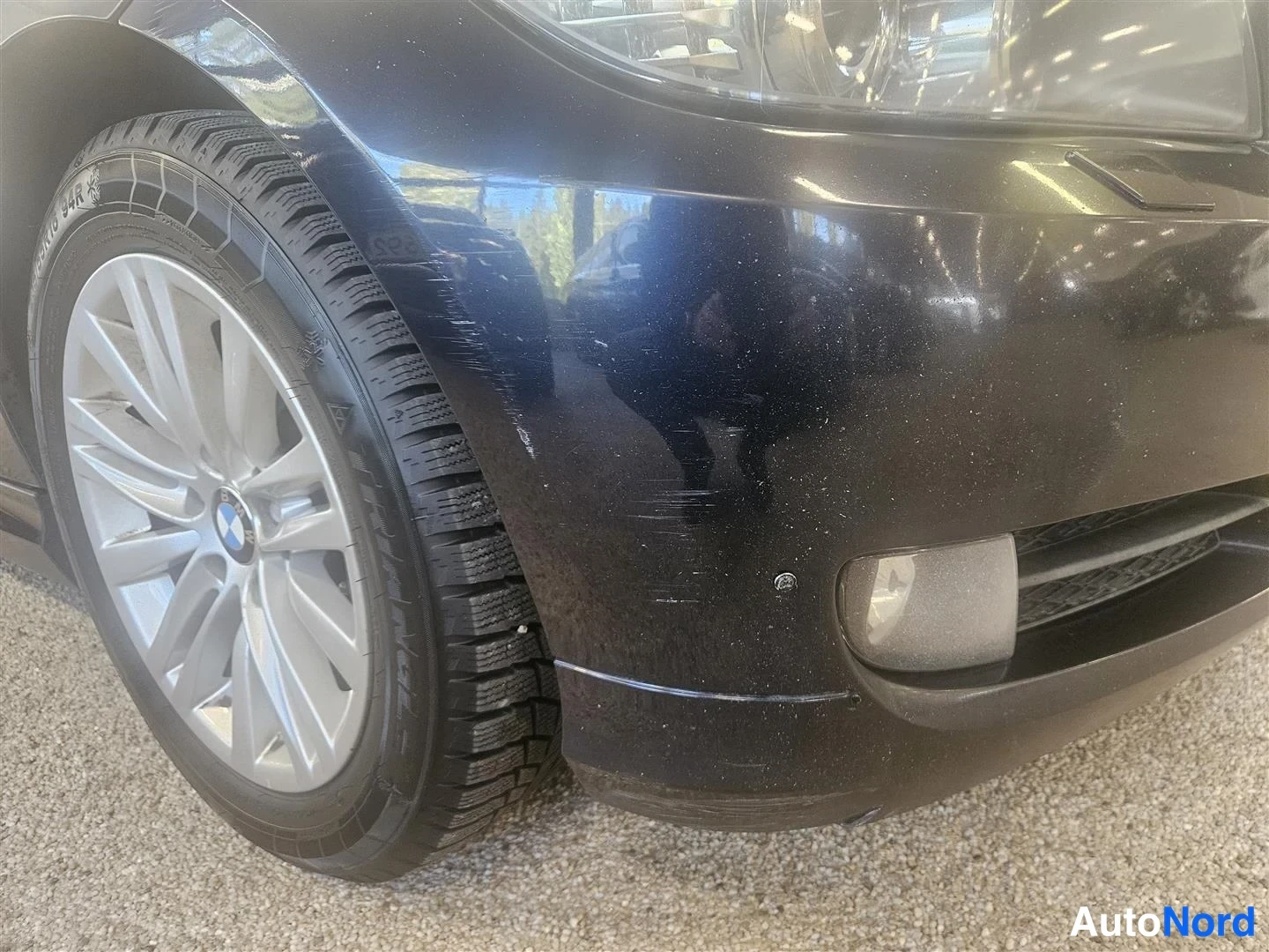 Yksityiskohta: Lähikuva auton oikeasta etukulmasta: musta etupuskuri ja etupyörä hopeisella vanemalla, jossa BMW:n merkki; puskurin pinnassa hierrareita ja naarmuja.
