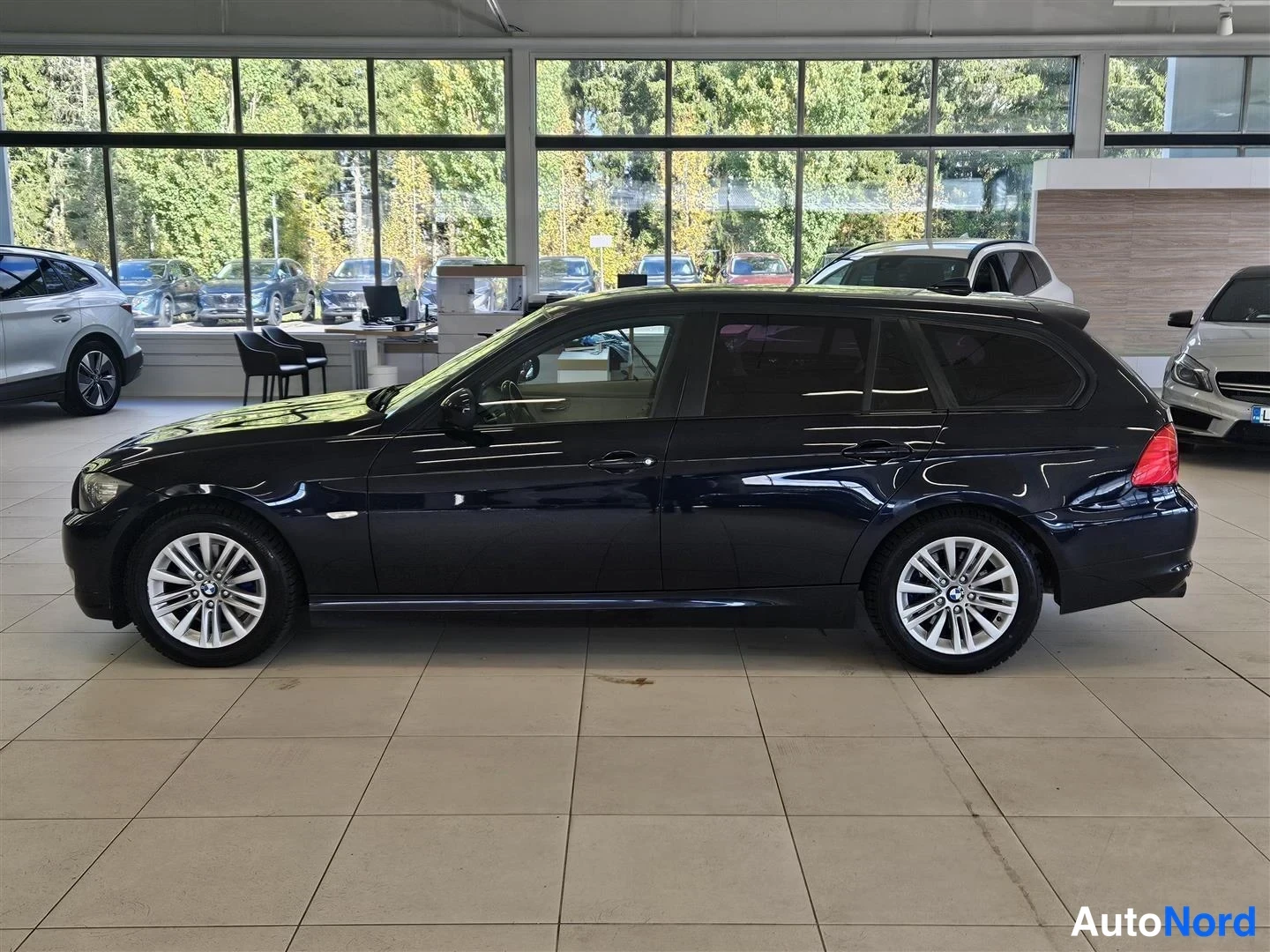Sivulta: Tumma BMW-farmari sivusta vasemmalta, auton kokoina siluetti showroomissa.