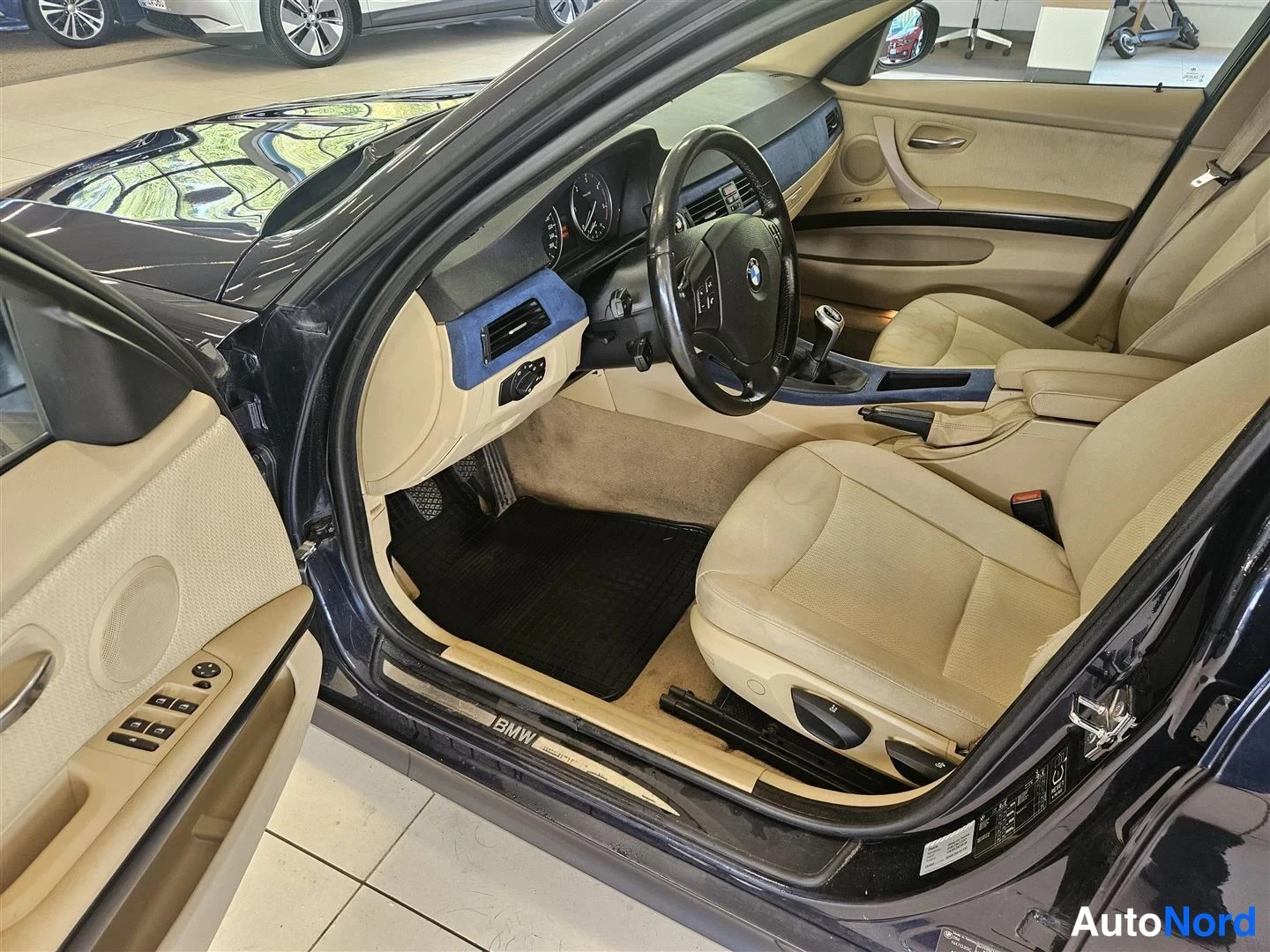 Muu: Avoin kuljettajan ovi ja etuistuimet: beigea sisustaa, BMW-ohjauspyörä, käsivalpytela ja musta matto etupohjassa.