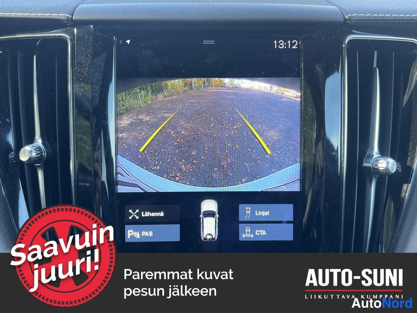Muu: Kojelaudan keskinäyttö, jolla näkyy peruutuskameran kuva parkkipaikasta ja ohjausviivat. Ruudussa painikkeet 'Lähennä', 'Linjat', 'PAS' ja 'CTA'. Kello 13:12.