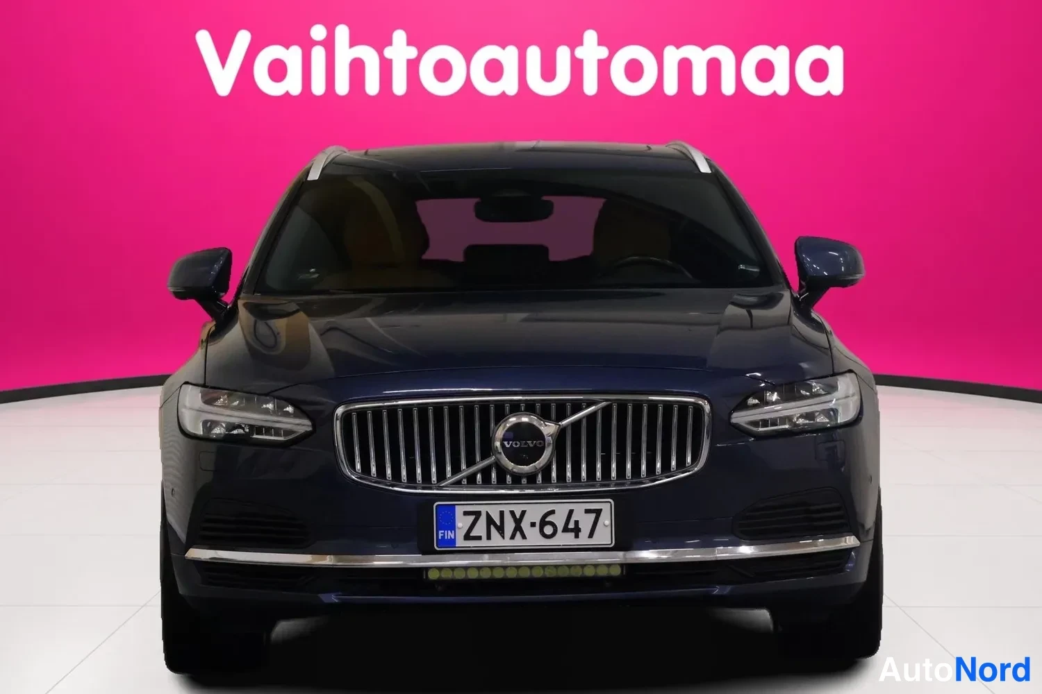Edestä: Volvo V90 -asema-auton etunäkymä tummansinisellä korilla, rekisteri, pinkkitaustainen studiokuva.